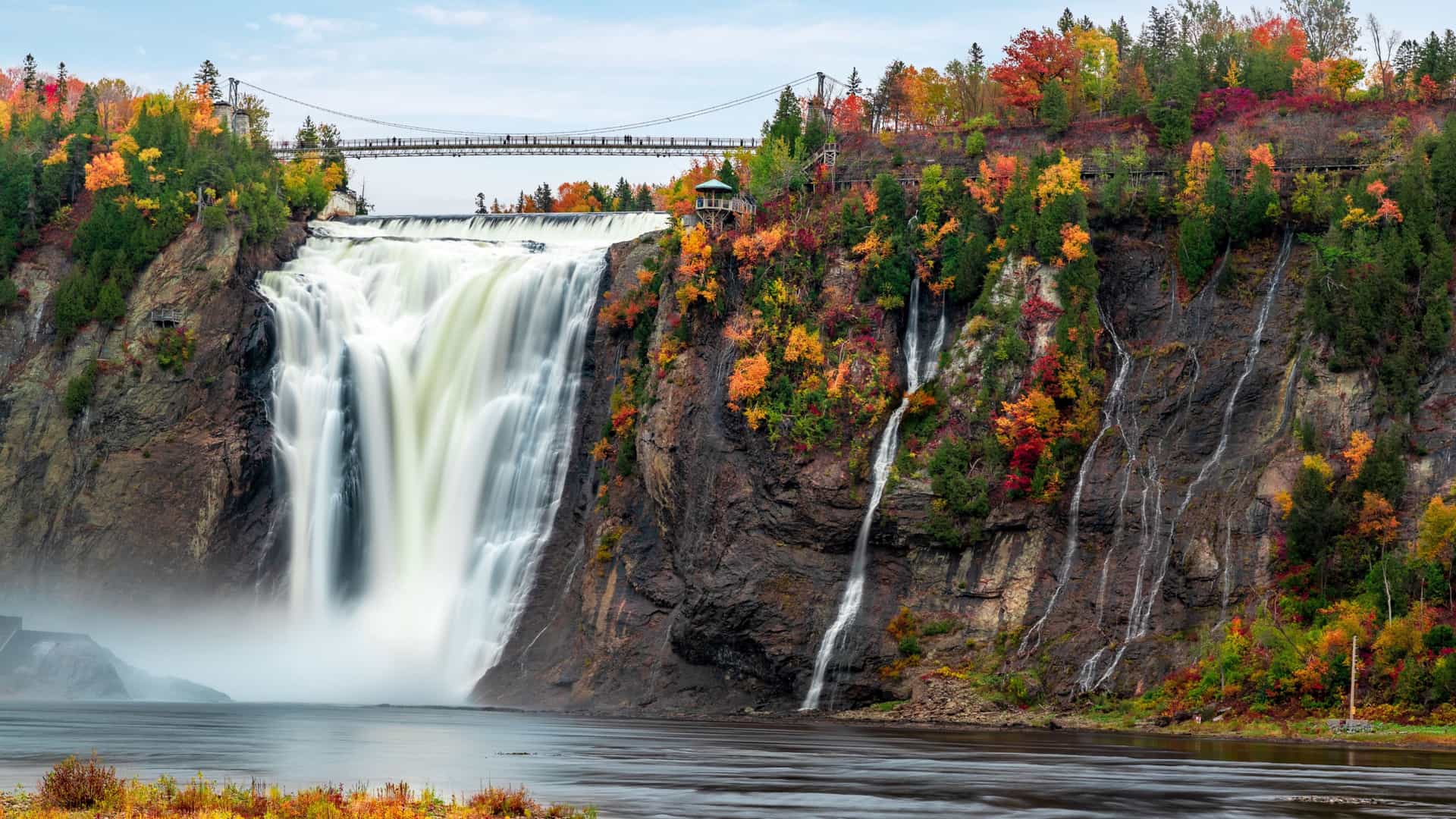 A massive waterfall cascades down rocky cliffs surrounded by vibrant autumn foliage, with a pedestrian suspension bridge spanning the top under an overcast sky.