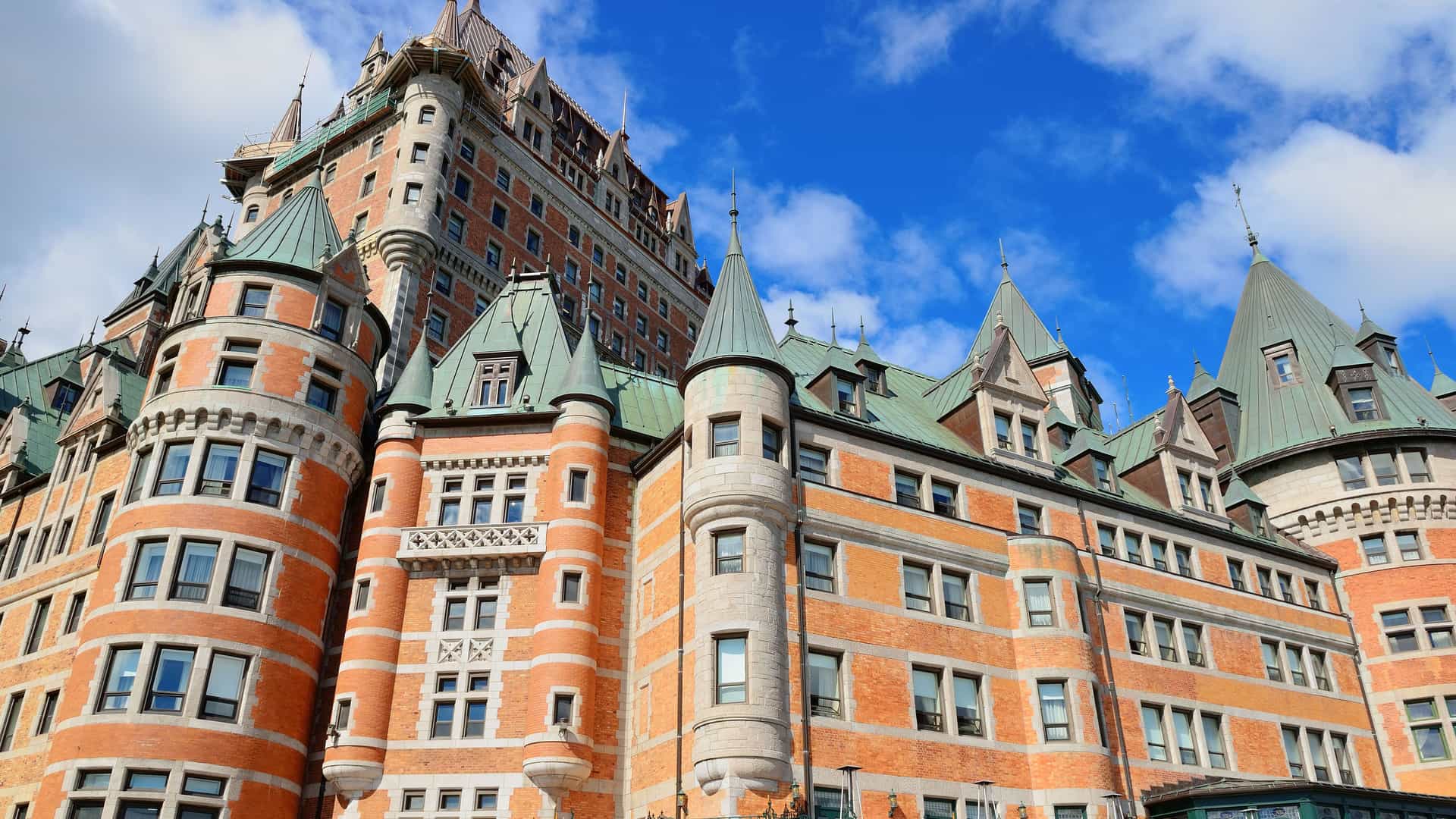 An impressive view of the Fairmont Le Château Frontenac in Quebec City, Canada. The grand hotel is built in a châteauesque style, with prominent red brick facades, multiple towers, and distinctive green copper roofs with sharp spires. The intricate architectural details, including dormer windows and ornate trim, are visible. The sky is a vibrant blue with fluffy white clouds, highlighting the castle-like appearance of this famous landmark in the Canada and New England region.