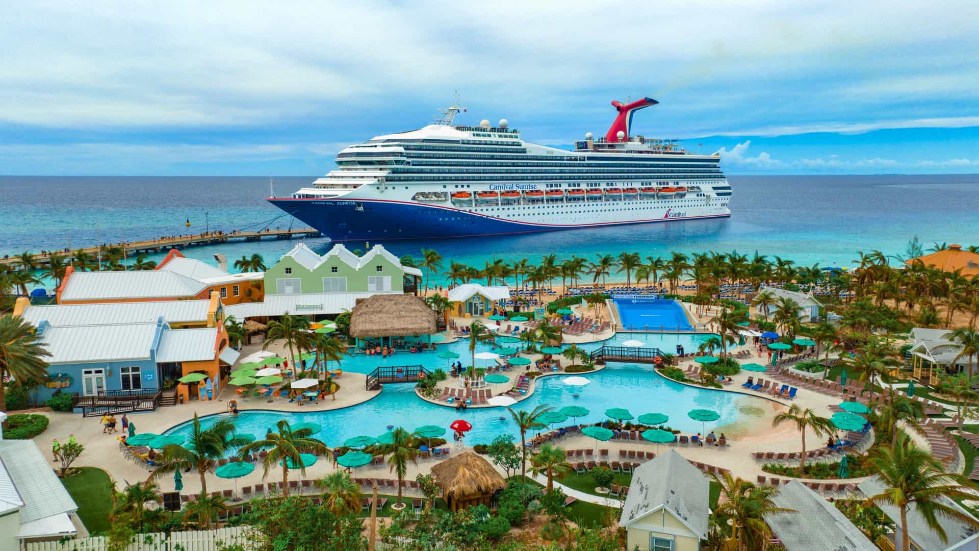 Aerial view of the vibrant Carnival Cruise Center in Grand Turk, Turks and Caicos, featuring a large pool area, palm trees, and a docked Carnival cruise ship, ideal for a fun cruise port day in the Bahamas.