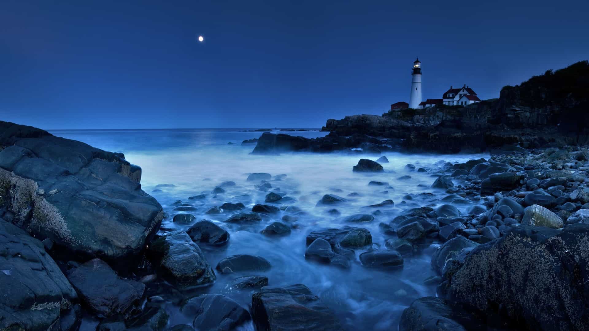 Moonlit Maine lighthouse on a rocky coastline at dusk—scenic stop on Carnival Cruises Canada & New England itinerary.