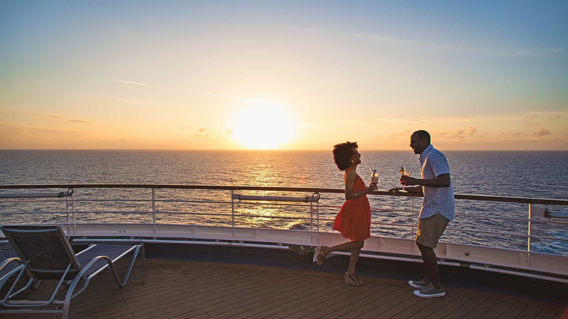 A couple enjoys tropical drinks on a Carnival cruise ship deck at sunset over the ocean, creating a romantic atmosphere, ideal for a Trans-Atlantic voyage.