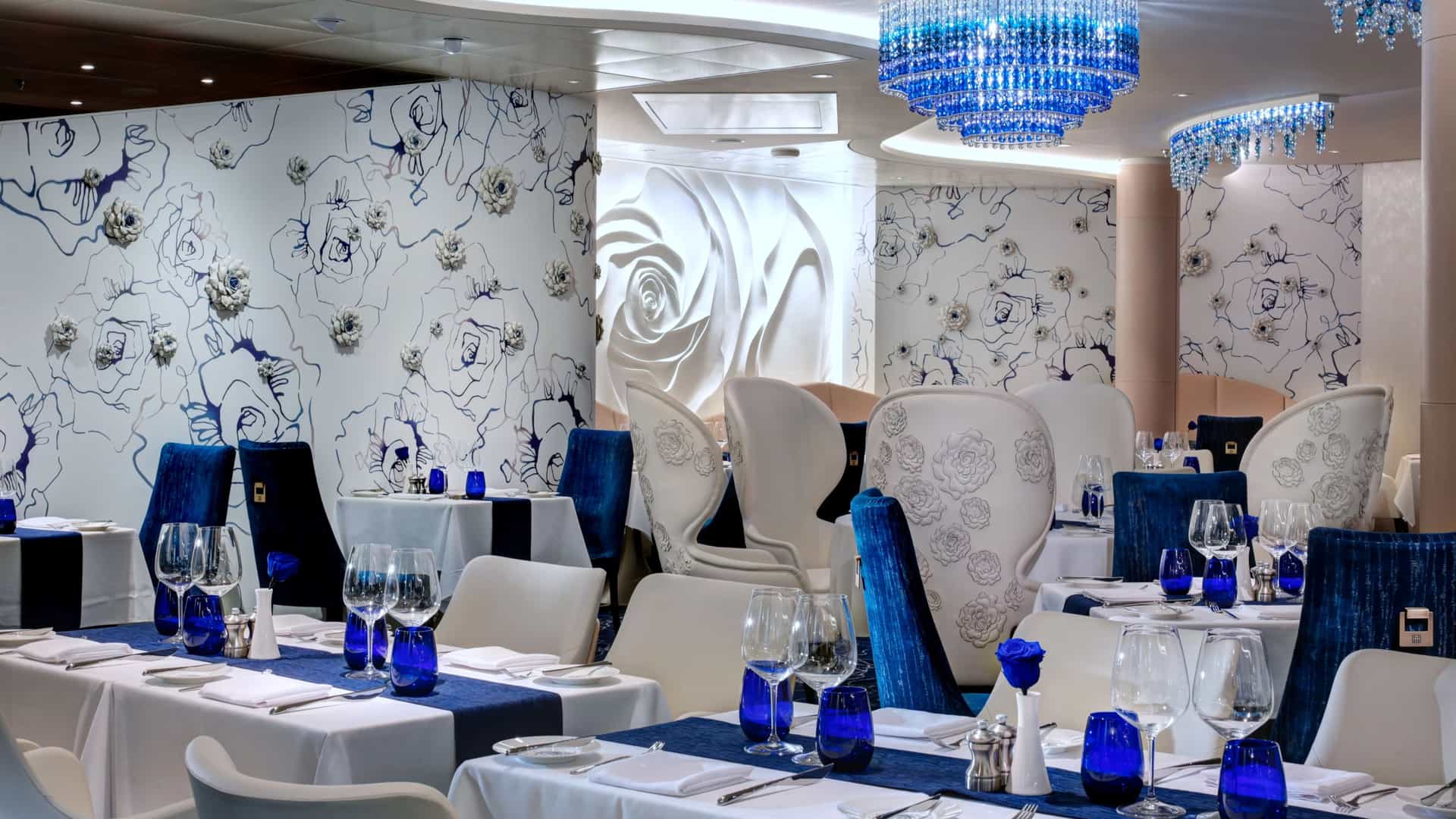 An elegant upscale restaurant interior featuring white-clothed tables with blue runners, blue glassware, and striking blue crystal chandeliers.