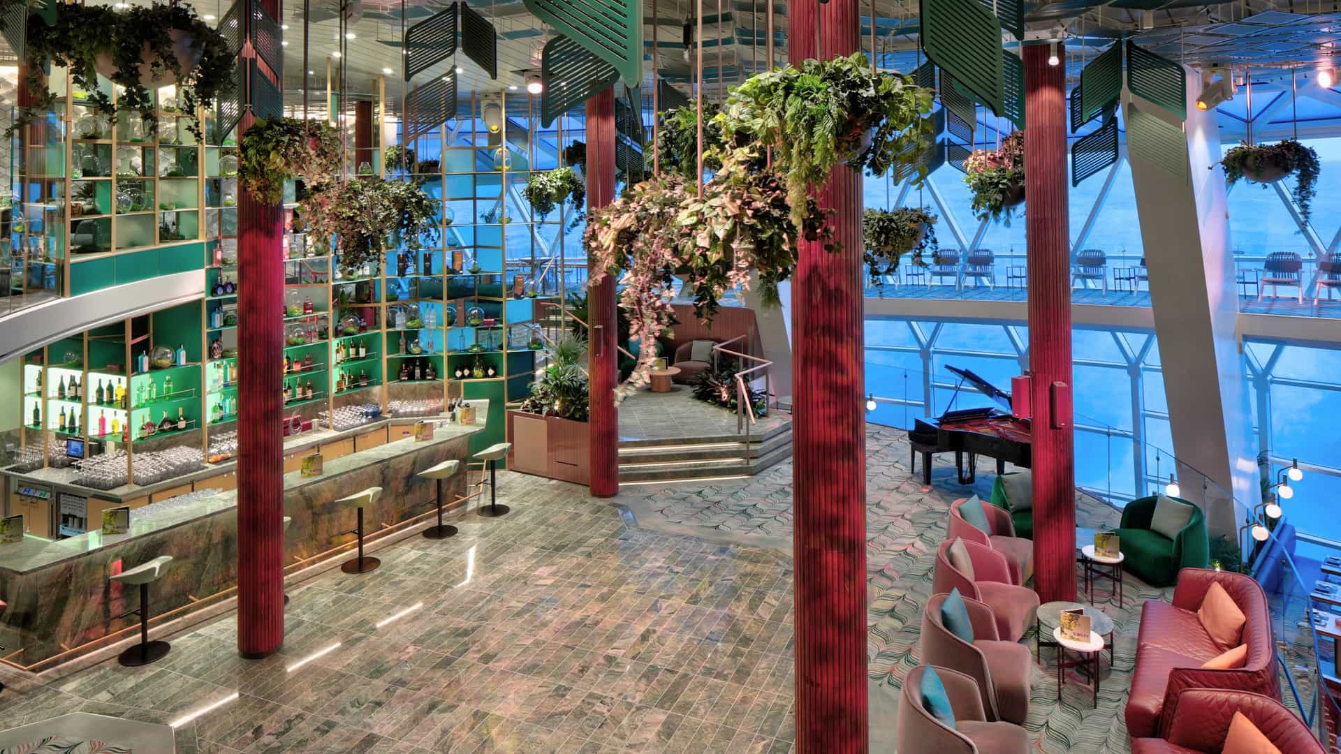 A lush, multi-level indoor lounge filled with hanging plants, pink and red seating, a bar, a grand piano, and expansive panoramic ocean-view windows.