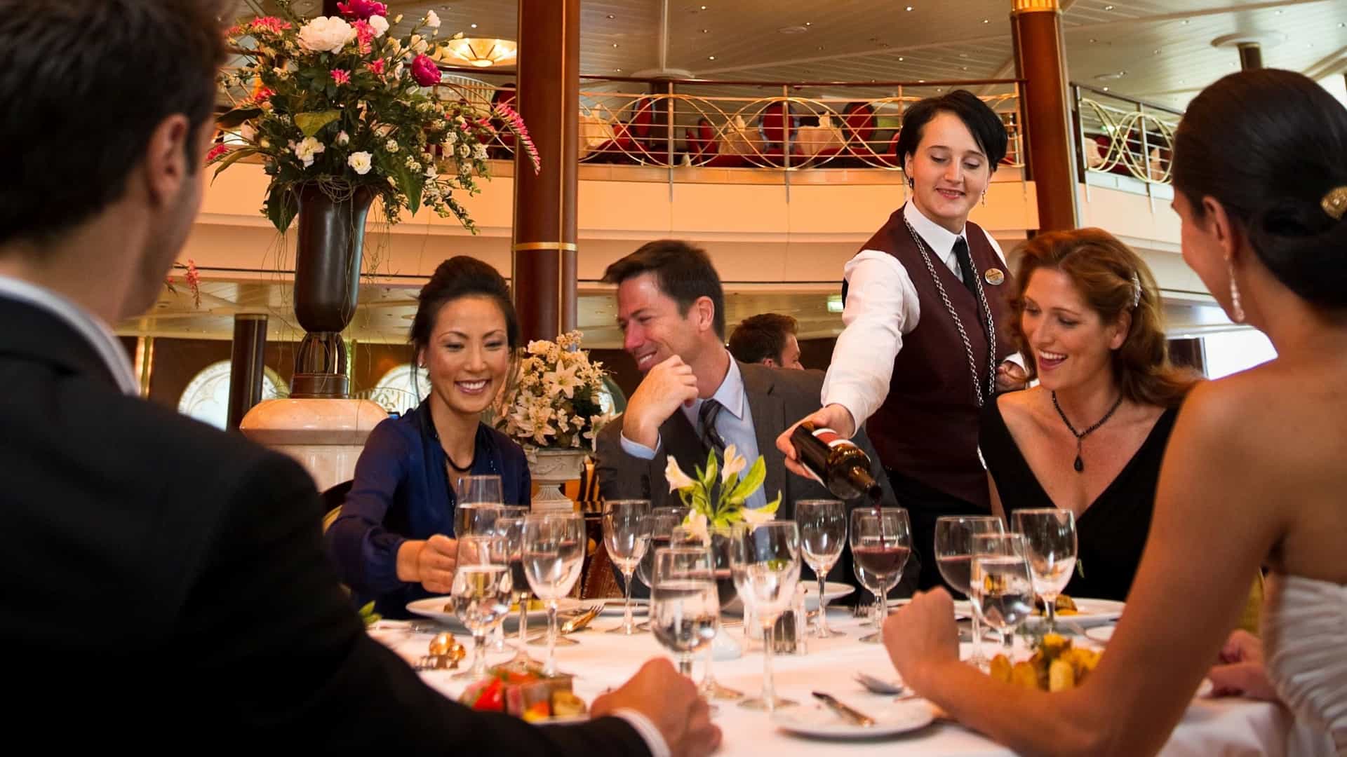 A smiling server pours wine for a group of formally dressed guests enjoying a fine dining experience at an upscale specialty restaurant aboard Celebrity Cruises.