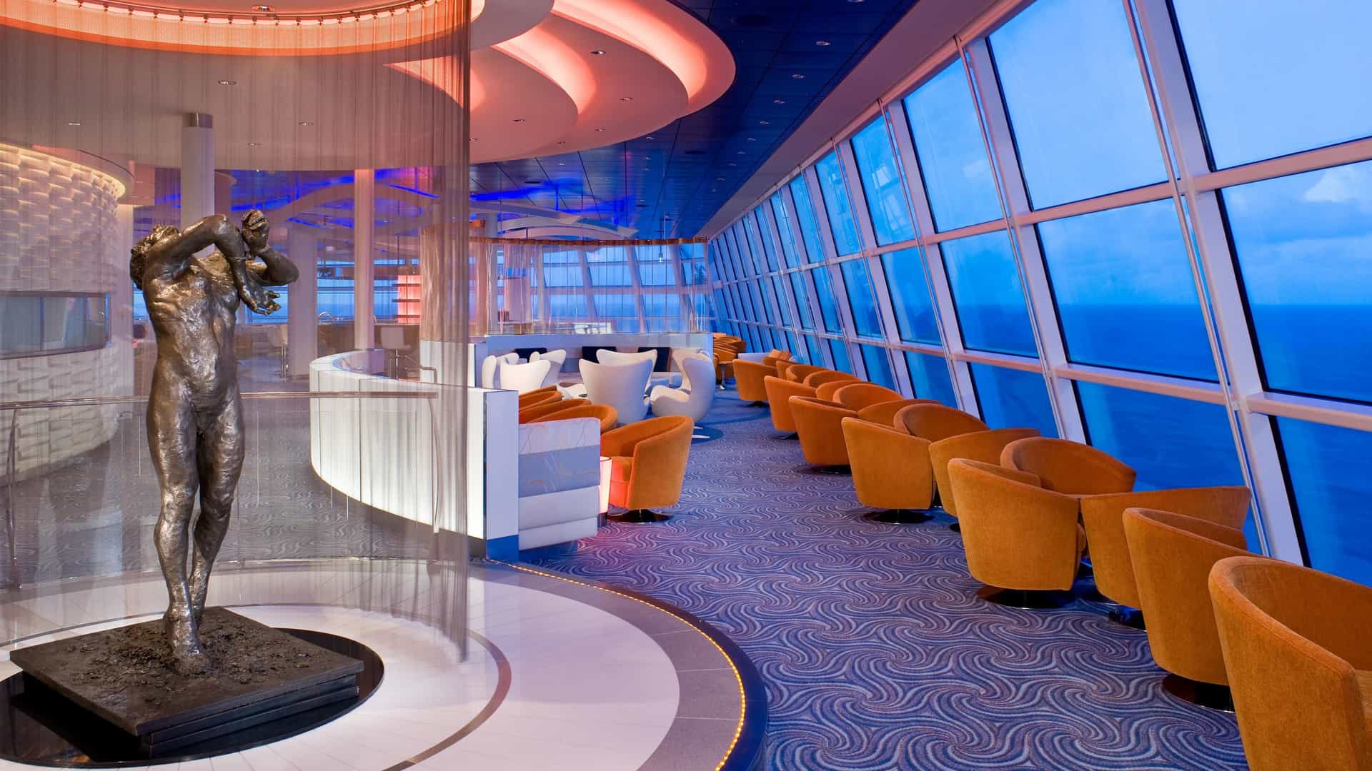An upscale indoor lounge featuring floor-to-ceiling panoramic ocean windows, rows of orange armchairs, and a prominent bronze sculpture in the foreground.