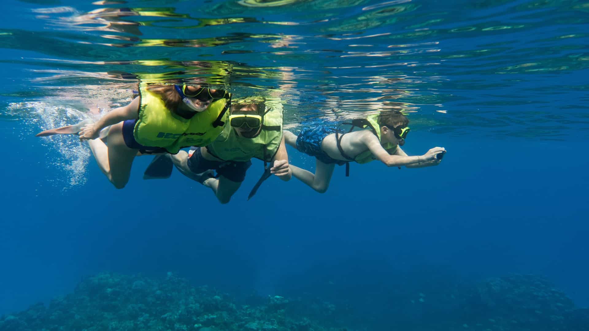Three people snorkeling over a coral reef in Grand Cayman’s turquoise waters on a Celebrity Cruises Western Caribbean excursion.