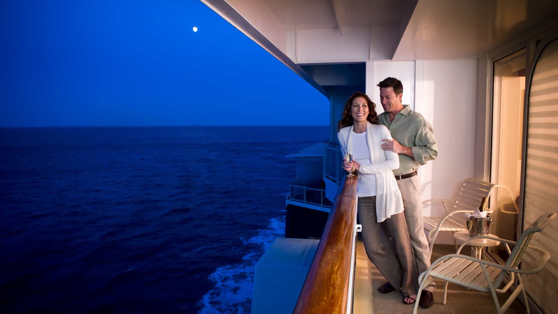 Couple on a Celebrity ship balcony at night, gazing at the moonlit ocean during a Trans-Atlantic sea journey.