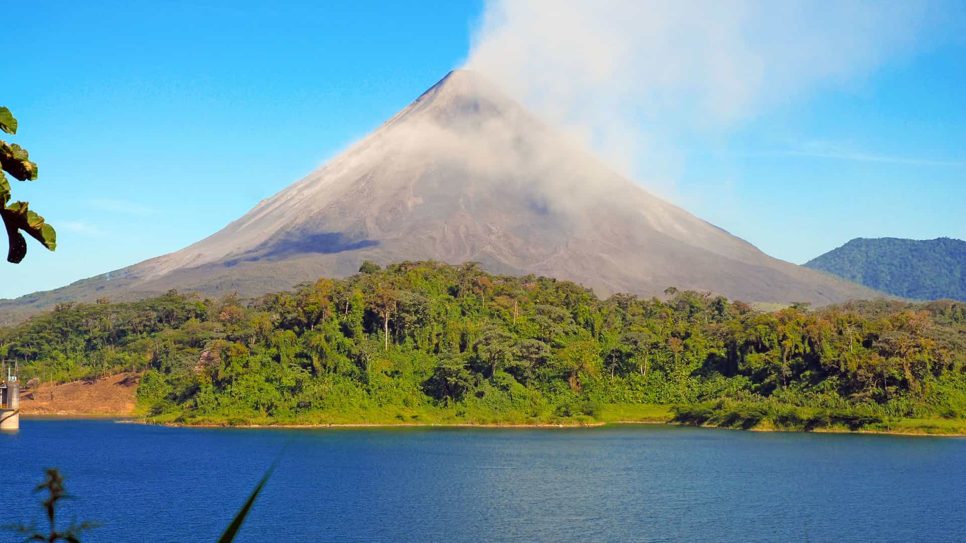 A stunning view of the Arenal Volcano, a cone-shaped stratovolcano in Costa Rica, rising prominently under a clear blue sky. A plume of white steam drifts from the peak and across its left side. The volcano's base is covered in a dense, green forest. In the foreground, a large, calm lake reflects the sky and the surrounding landscape. The image captures the powerful and majestic scenery of the Arenal region in Costa Rica.