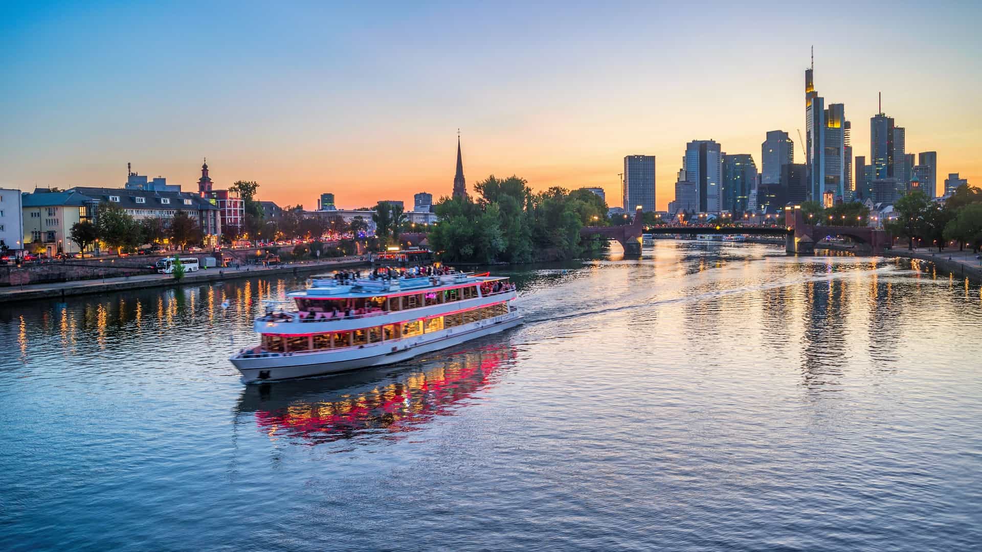A beautiful evening view over the Main River in Frankfurt am Main, Germany, showing a brightly lit touristic cruise ship passing under a bridge, with the illuminated city skyline in the background.