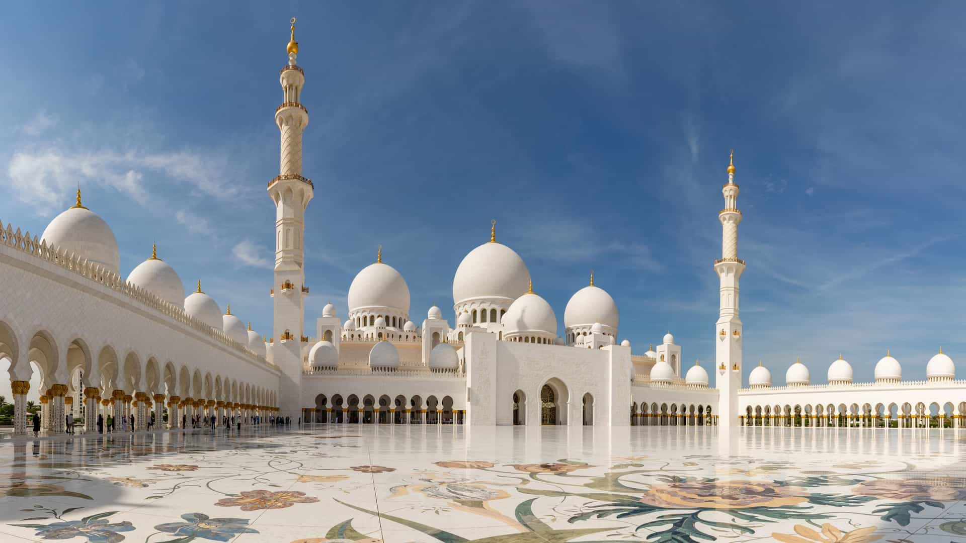 A serene, symmetrical view of the white marble Sahan courtyard of the Sheikh Zayed Grand Mosque in Abu Dhabi, United Arab Emirates, featuring a complex floral mosaic on the pavement, surrounded by ornate columns and arches, under a clear blue sky.
