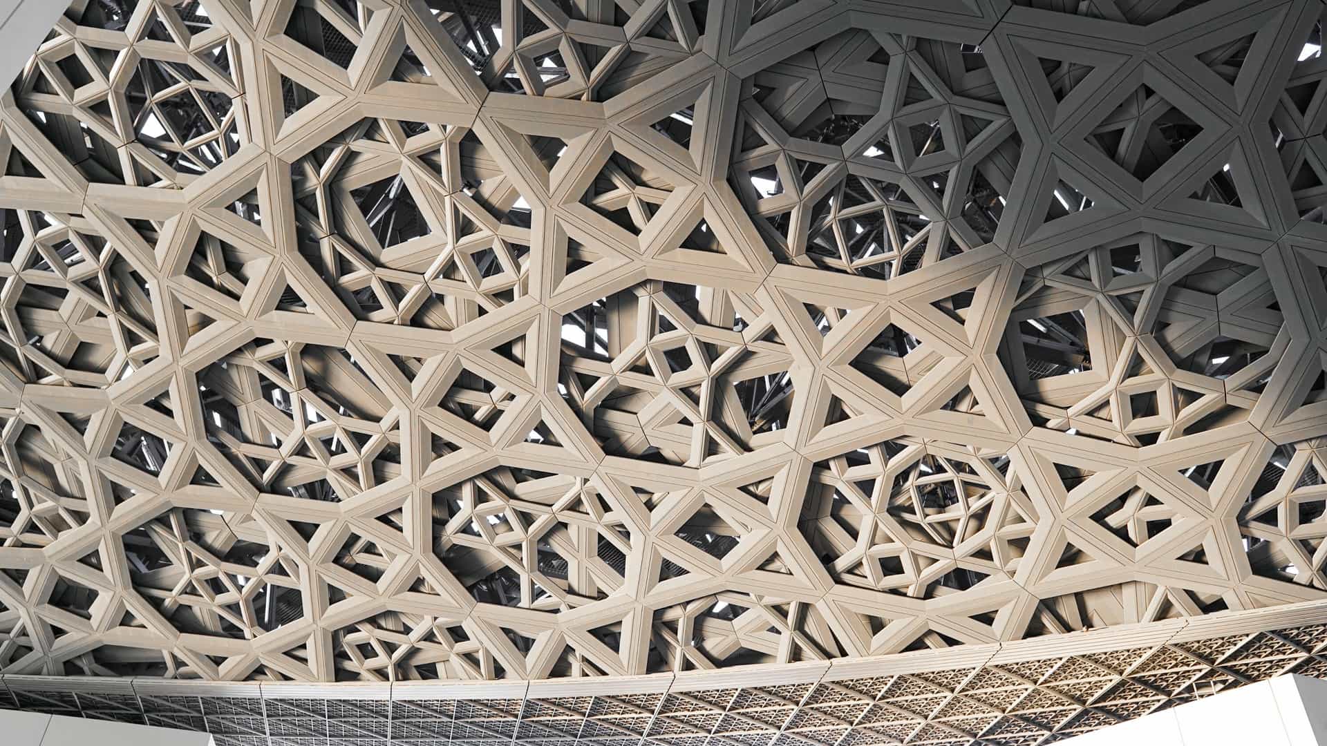 Exterior of the Louvre Abu Dhabi, UAE, featuring the museum's iconic, massive perforated dome roof that creates a 'rain of light' effect over the central complex and surrounding water on Saadiyat Island.