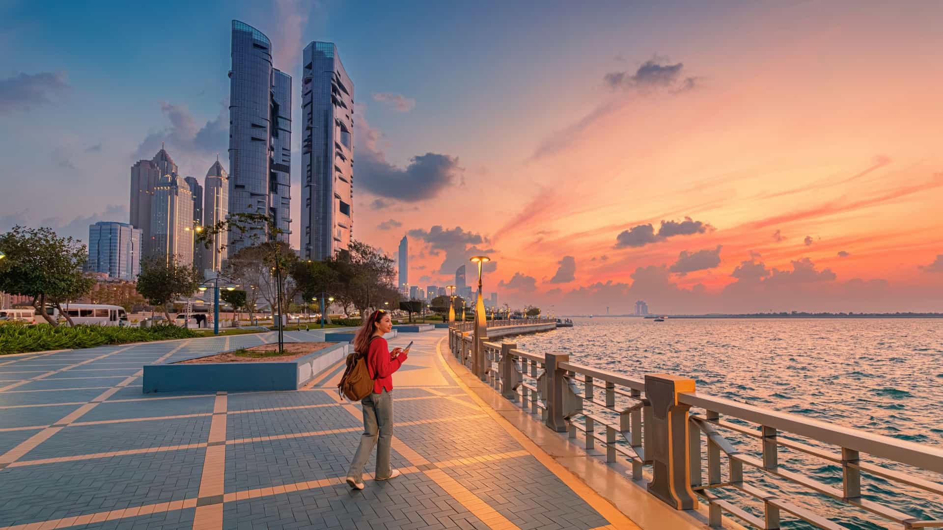 A person walking along the Abu Dhabi Corniche waterfront promenade, with green grass and palm trees in the foreground and a line of modern skyscrapers and a light-colored mosque in the background, all under a bright blue sky.