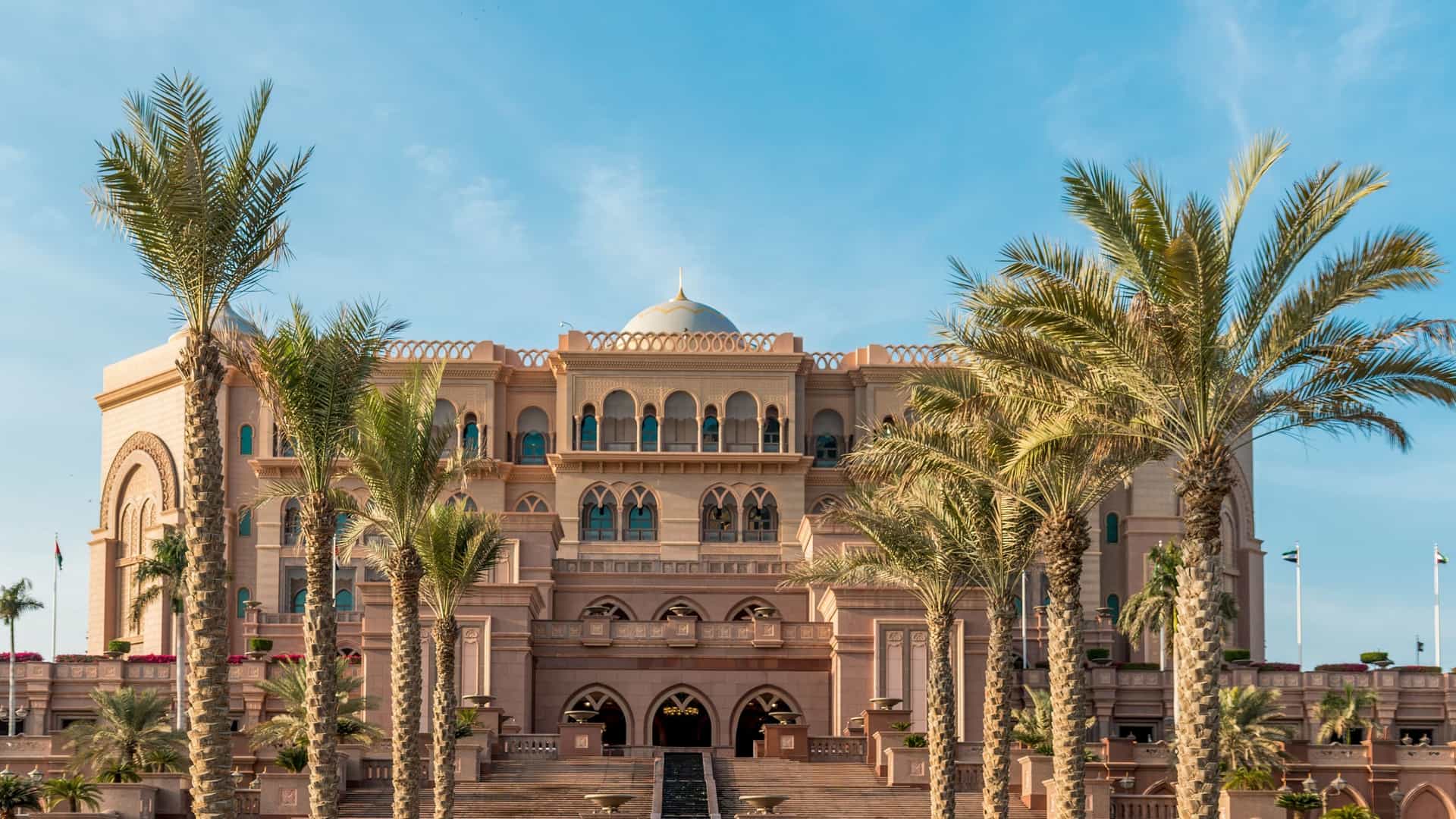 A serene, symmetrical view of the white marble Sahan courtyard of the Sheikh Zayed Grand Mosque in Abu Dhabi, United Arab Emirates, featuring a complex floral mosaic on the pavement, surrounded by ornate columns and arches, under a clear blue sky.