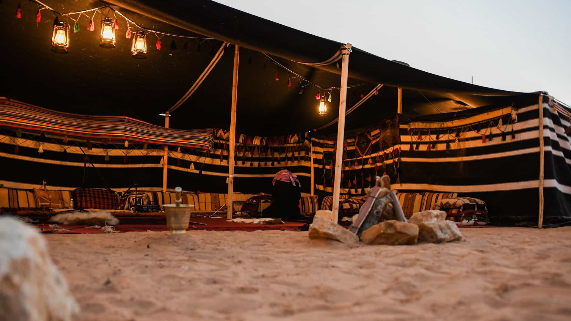 A view of a traditional Arabian desert scene, featuring a black Bedouin tent on the left and a traditional mud-brick house on the right, with a wooden door. The scene is set on a desert landscape with small shrubs and a clear sky, exposing the Arab heritage in Saudi Arabia.