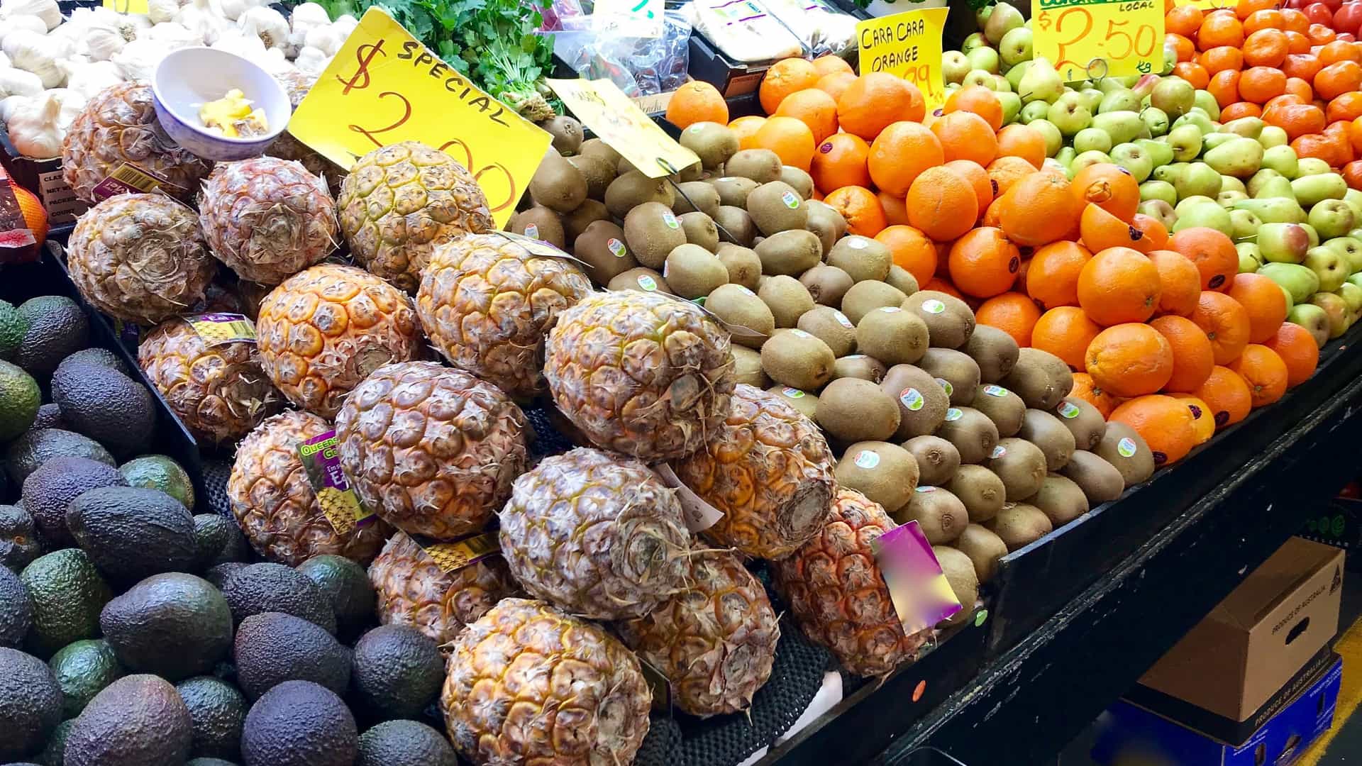 A vibrant, colorful display of fresh, seasonal fruit for sale at a stall inside the Adelaide Central Market, South Australia, featuring pineapples, mandarins, kiwifruit, and pears.