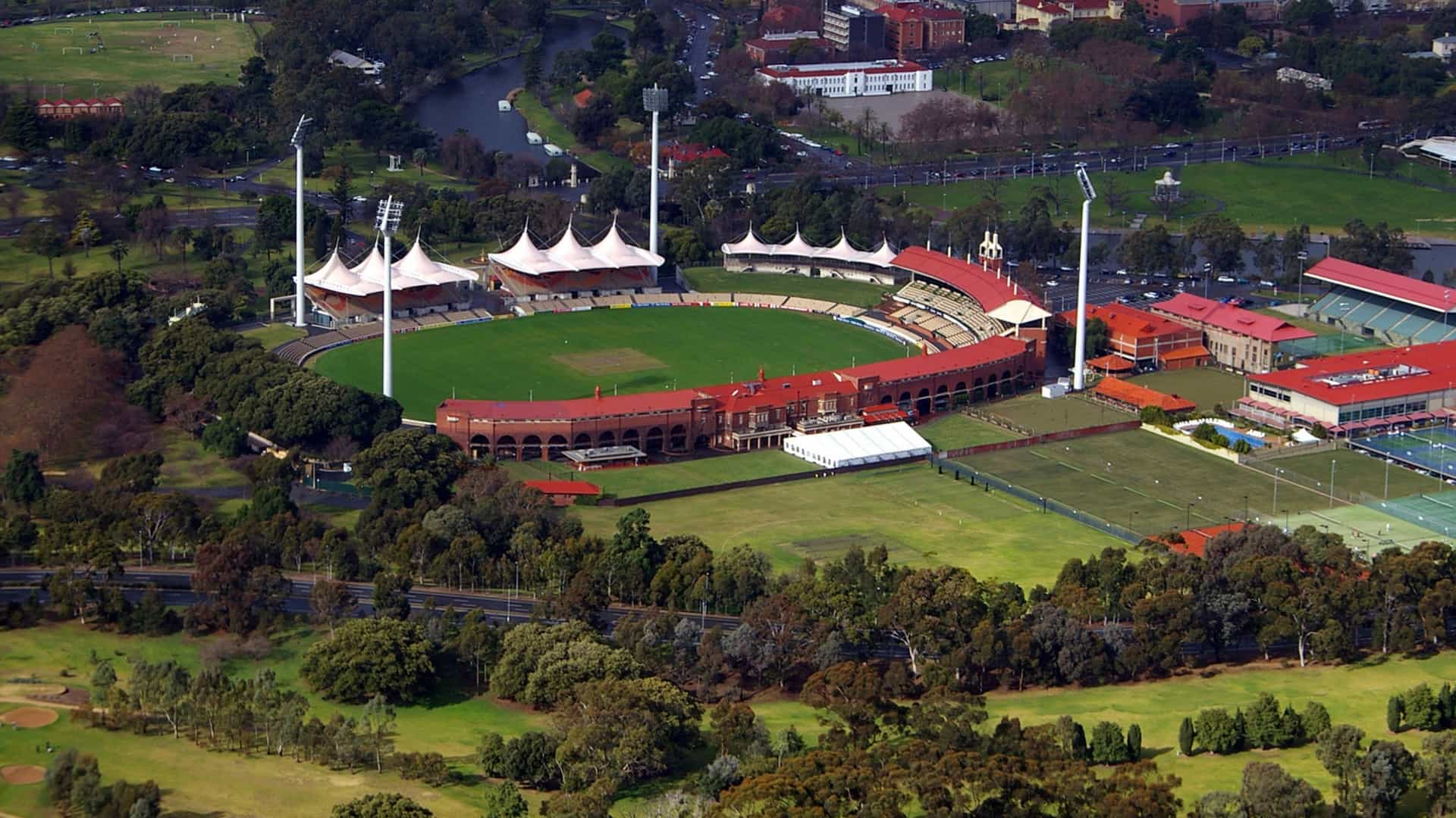 An aerial, high-angle view of the Adelaide Oval cricket and sports stadium in Adelaide, South Australia. The lush green oval and pitch are surrounded by seating stands, and the stadium is set within a large green parkland next to a river. In the background, the modern skyscrapers of the Adelaide city skyline rise under a bright, sunny sky.