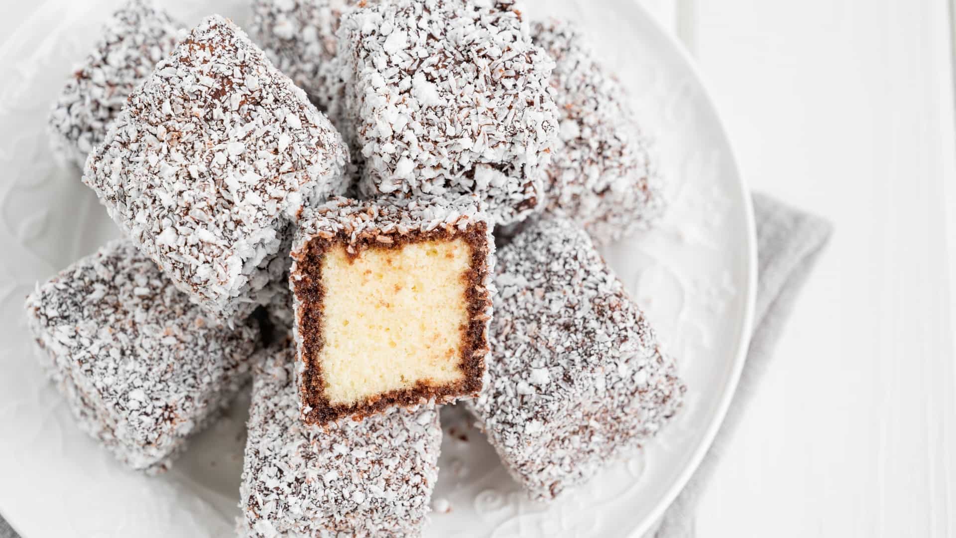 A close-up view of several traditional Australian Lamington cakes, which are squares of fluffy sponge cake completely coated in a dark chocolate icing and generously rolled in white, shredded desiccated coconut.