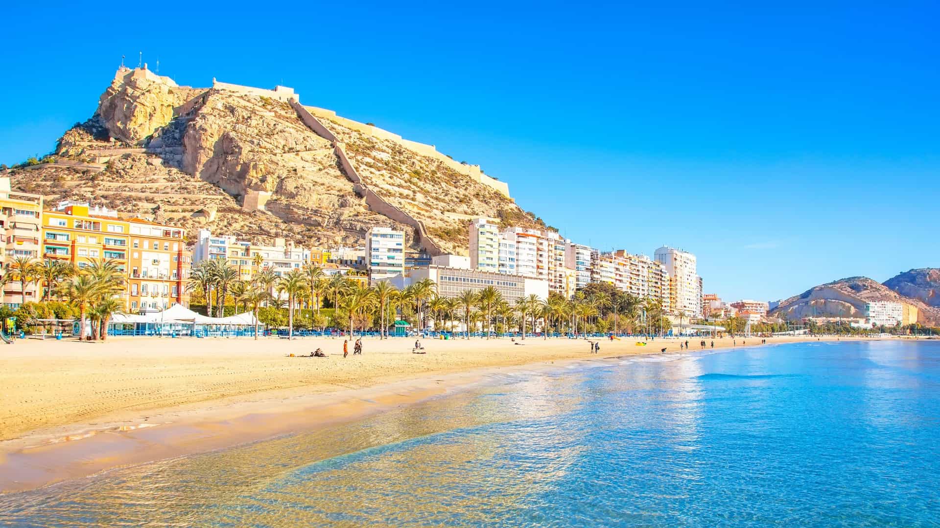 Postiguet Beach (Playa del Postiguet) in the resort town of Alicante, Spain, showcasing its sandy crescent beach, palm trees, and the clear Mediterranean Sea.