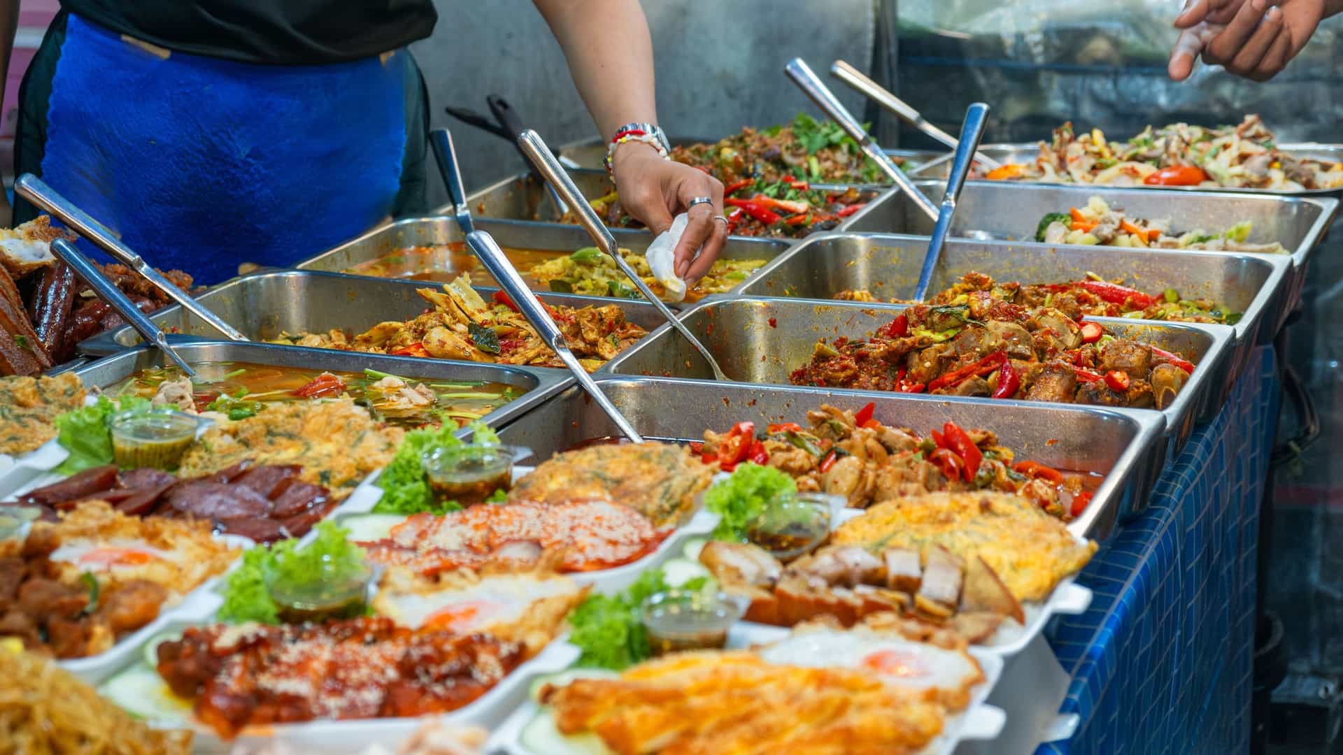 A vendor serves various prepared Thai dishes, including curries and fried eggs, from a bustling street food stall in Bangkok.