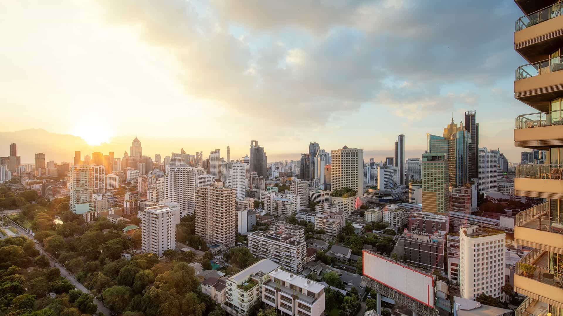 An elevated view of the Bangkok city skyline at sunset from a high-rise rooftop bar. The sky is painted with vibrant orange, yellow, and purple hues as the sun dips below the horizon, illuminating the numerous skyscrapers and city lights across the dense urban landscape.