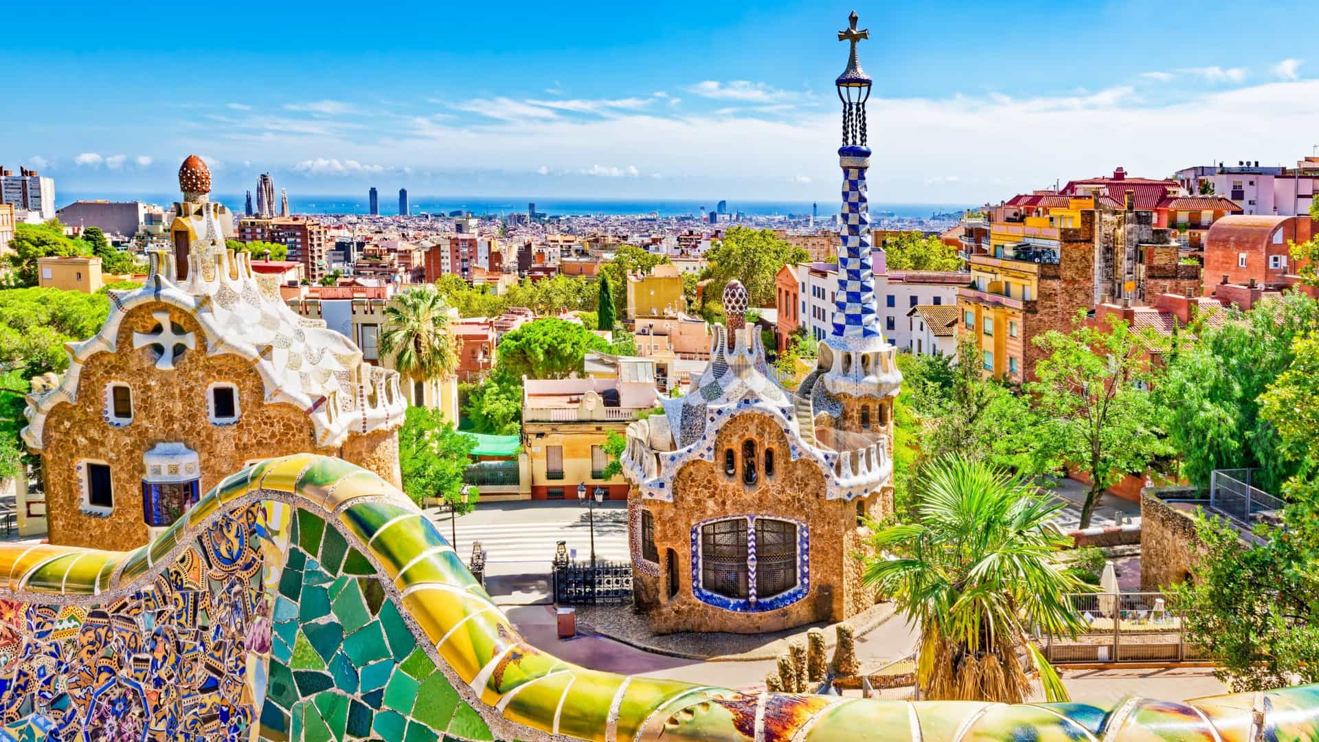 A detail shot of the architectural elements at Park Güell in Barcelona, Spain. The image features a vibrant, curving bench decorated with multi-colored mosaics in Gaudí's unique trencadís style, with a view of the park's natural surroundings in the background.