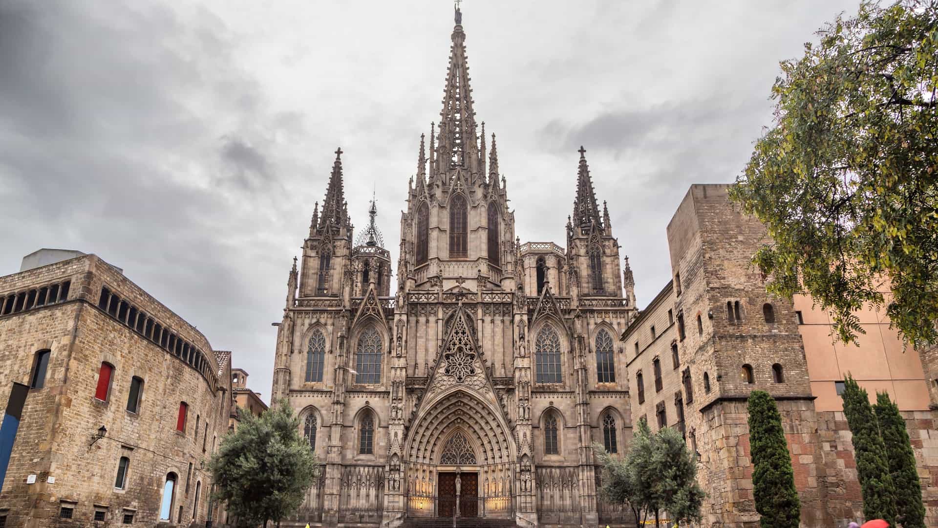 A view of the Barcelona Cathedral, officially known as the Cathedral of the Holy Cross and Saint Eulalia, on a rainy morning. The image highlights the detailed neo-Gothic façade of the cathedral, with wet pavement reflecting the architectural details in the foreground. The scene is located in the Gothic Quarter of Barcelona, Spain.