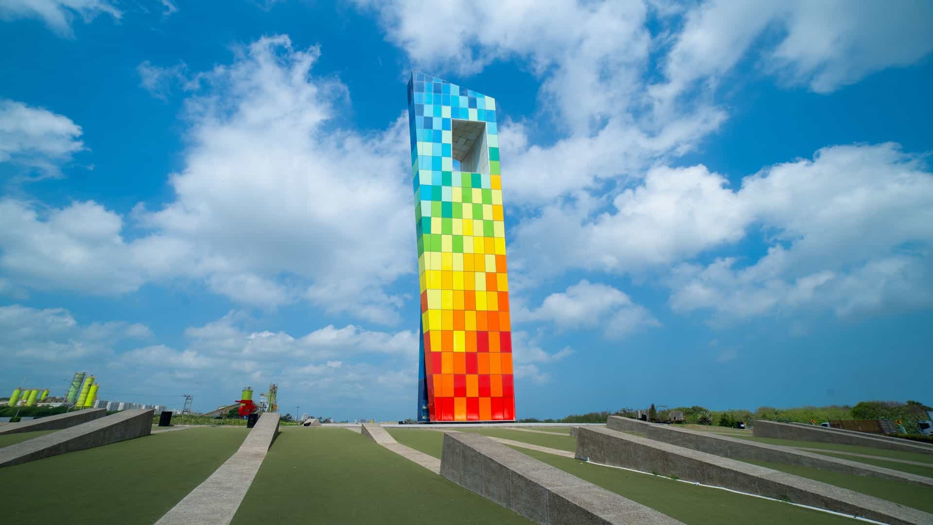 The "Ventana al Mundo" (Window to the World) Monument in Barranquilla, Colombia, on a clear day. The modern, towering structure consists of two large, colorful vertical frames made of glass and steel (one red, one blue), forming a window-like arch. It is situated in a roundabout, with a blue sky visible through the frame. The monument was built to commemorate the 2018 Central American and Caribbean Games.