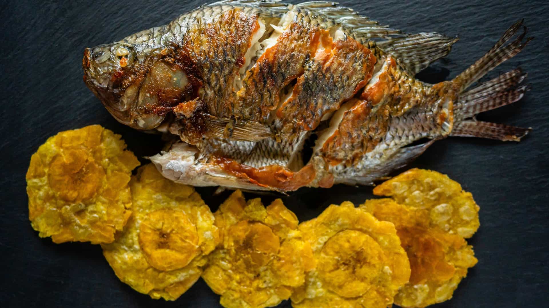 A top-down view of a plate featuring a delicious fried Mojarra fish (Mojarra Frita), a traditional Latin American dish. The whole, crispy, golden-brown fish is served alongside several round, golden patacones (twice-fried plantain slices) on a dark, rustic plate or black stone surface. The dish is commonly served on the Caribbean coast of Colombia.