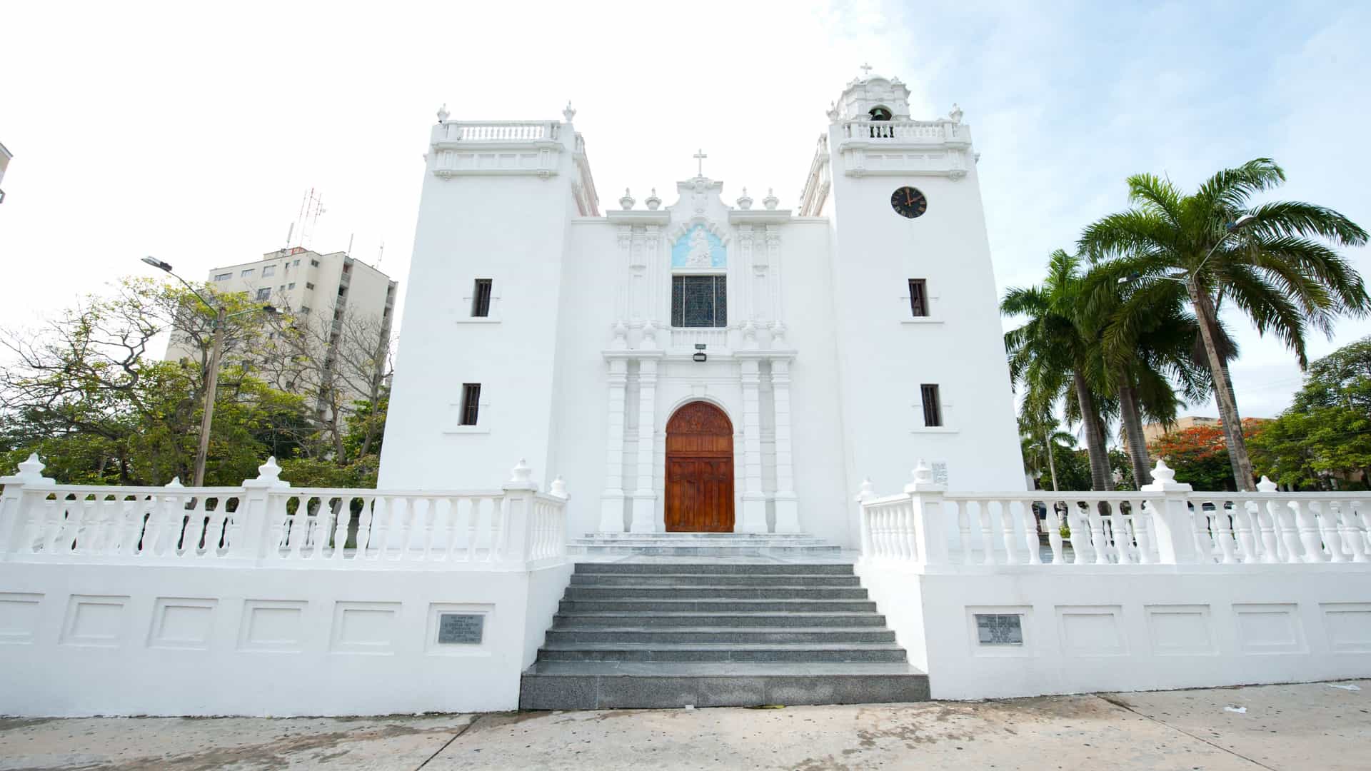 A view of the white facade of the Immaculate Conception Church (Iglesia de la Inmaculada Concepción) in the El Prado neighborhood of Barranquilla, Atlántico, Colombia. The church features two symmetrical bell towers and a wide set of steps leading up to the entrance, all set against a clear sky.