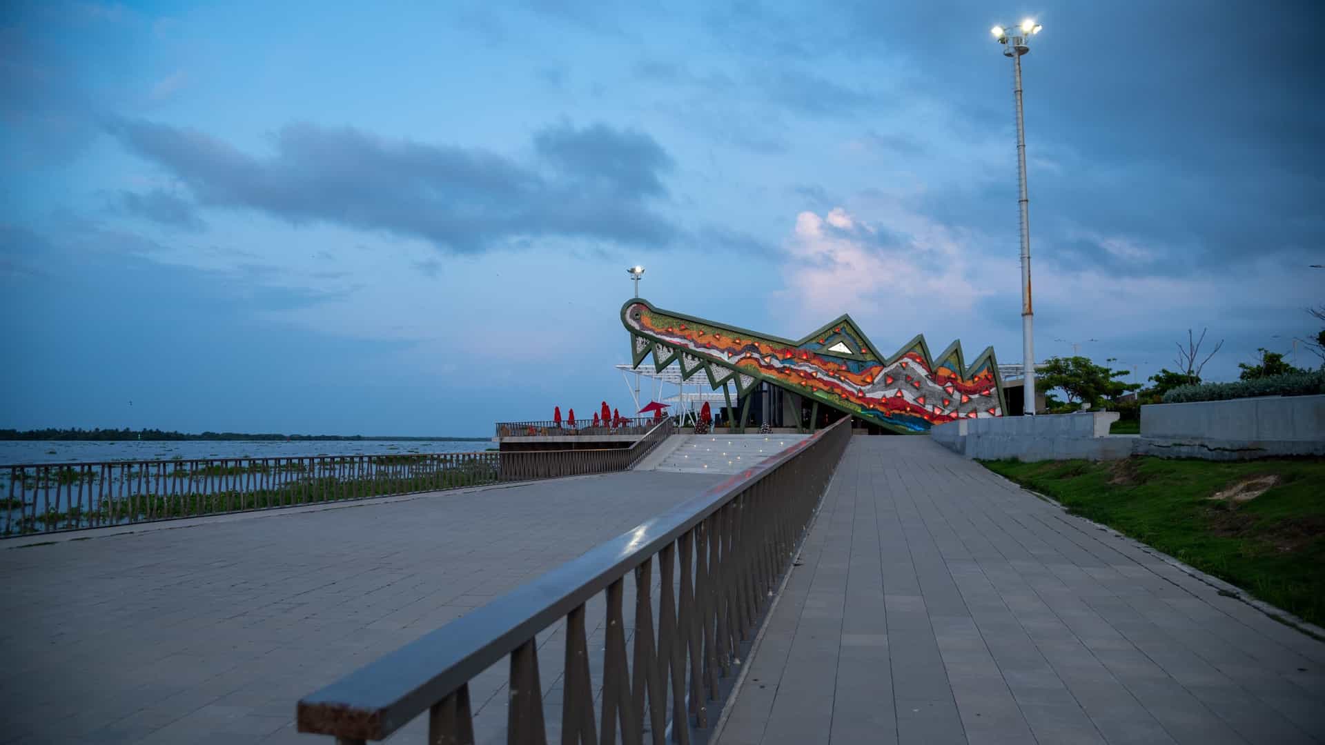A modern, vibrant night view of the "Caimán del Río" restaurant and bar complex on the Gran Malecón in Barranquilla, Colombia. The area is brightly lit with warm-toned string lights and spotlights, highlighting the contemporary architecture and creating a lively atmosphere alongside the river.