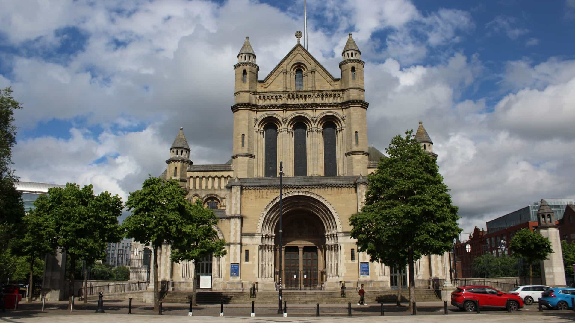  An exterior, daytime view of St. Anne's Cathedral (Belfast Cathedral) on Donegall Street in Belfast, Northern Ireland. The Romanesque style stone building, featuring a large arched entrance and detailed stonework, is topped by the tall, modern, stainless steel Spire of Hope rising into the blue summer sky.