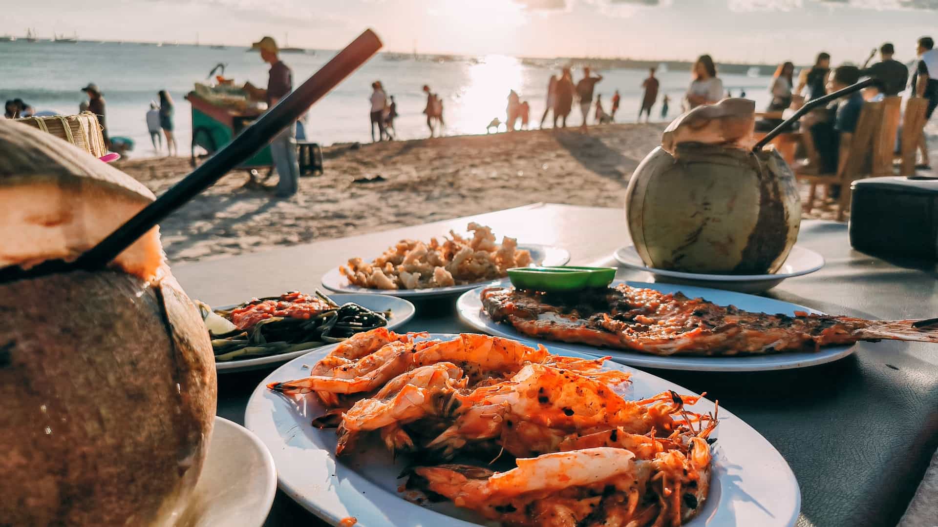  A close up view of a platter of freshly grilled Balinese seafood, including a whole fish and several large prawns (shrimp), served with rice and dipping sauces. The food is presented on a table on the sand of Jimbaran Beach, Bali, at sunset, with the blurred background showing the warm orange and pink glow of the sky over the ocean.