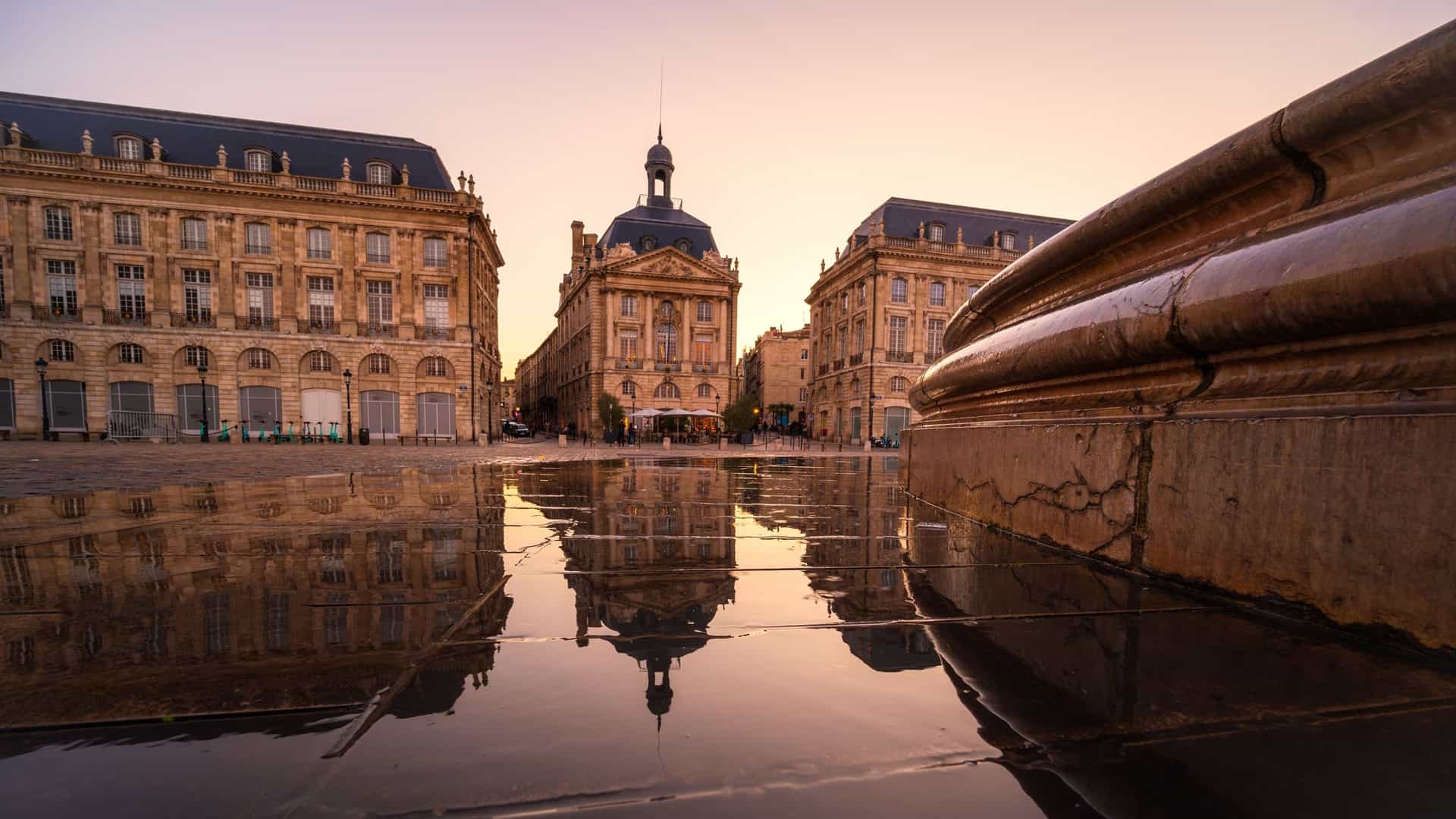  A scenic view of the Place de la Bourse in Bordeaux, France, at dusk. The 18th century neoclassical buildings, including the Palais de la Bourse and the former Hôtel des Fermes, are symmetrically arranged in a semi circle. In the foreground, the Miroir d'eau (Water Mirror) reflects the illuminated facades of the buildings, creating a stunning visual effect.