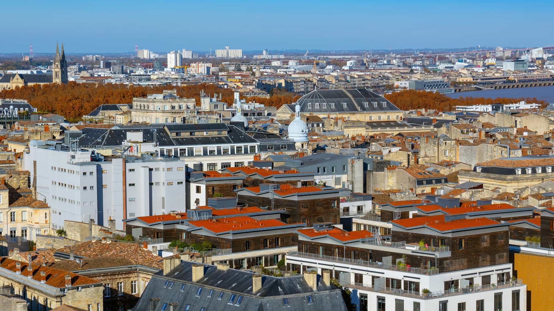  A panoramic elevated view of the central and northeastern parts of Bordeaux, France. The densely packed historic city is visible, characterized by a uniform field of light colored stone buildings with red tiled rooftops. The Garonne River flows through the background, with a modern cable stayed bridge (likely the Pont Jacques Chaban Delmas) visible crossing the water toward the upper right corner. The sky is bright blue with scattered white clouds.