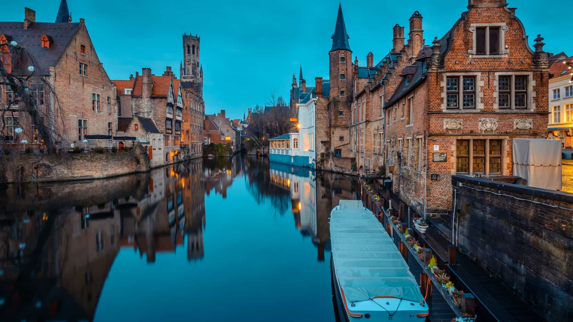 A picturesque view of Bruges, West Flanders, Belgium, featuring the historic cityscape with medieval architecture, a canal, and the Belfry of Bruges towering in the background.