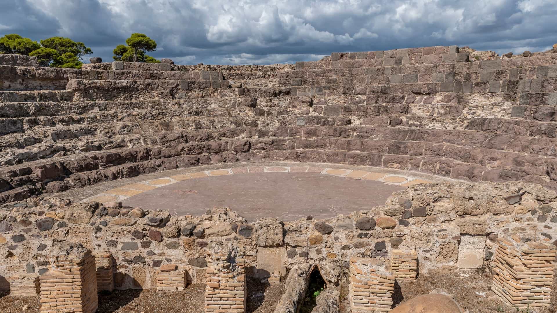 The ancient ruins of the Roman Theatre at Nora, Pula, Sardinia, Italy, situated by the sea, featuring the semi-circular stone seating tiers and the remaining structure of the stage area.