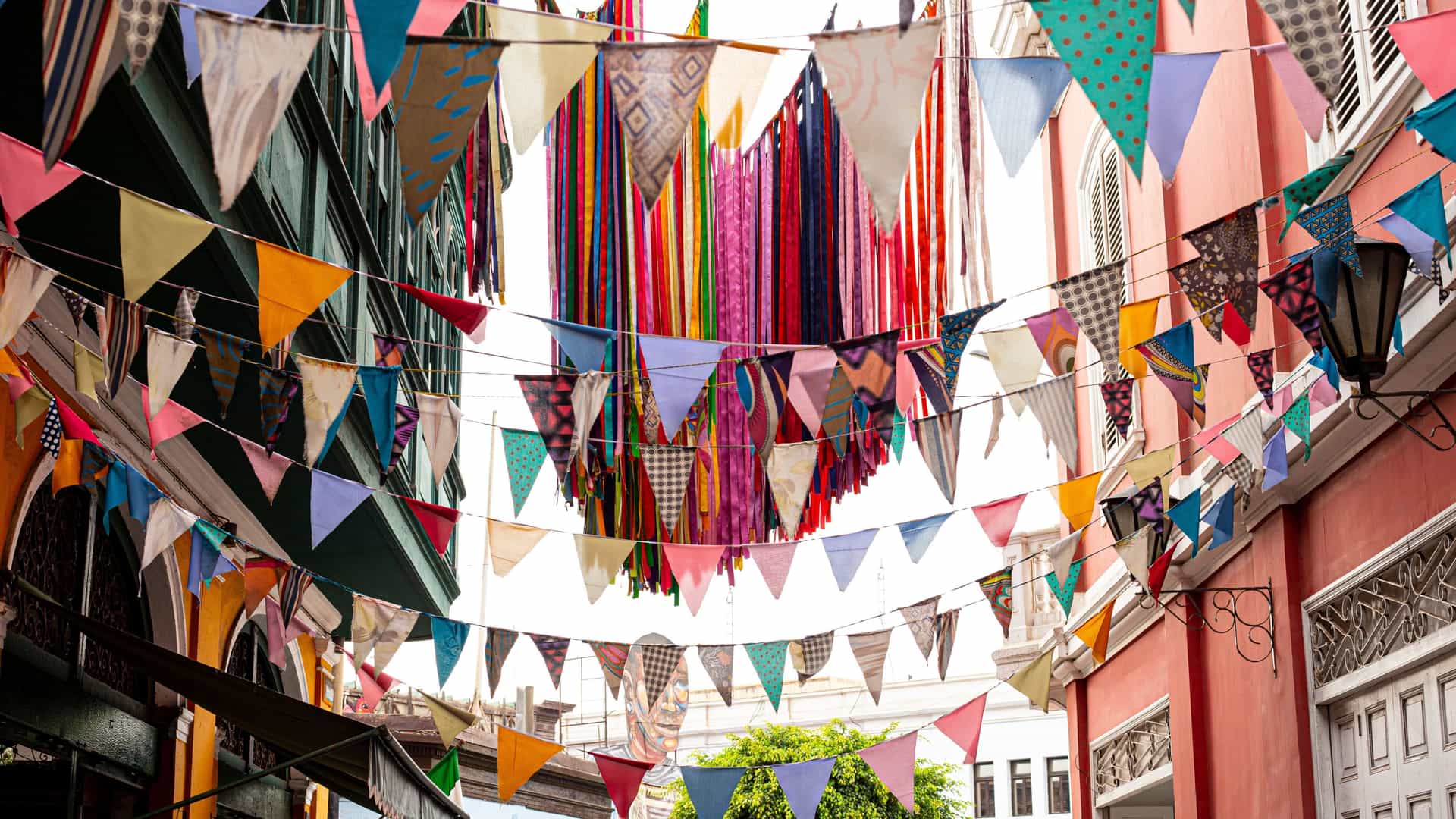 A vibrant, colorful array of paper chains hangs suspended across an alleyway or courtyard within the arts district of Monumental Callao, Peru. The chains, made of brightly colored paper links, create a festive canopy over the historic, colonial-era architecture, suggesting a celebration or artistic installation in this vibrant cultural area.