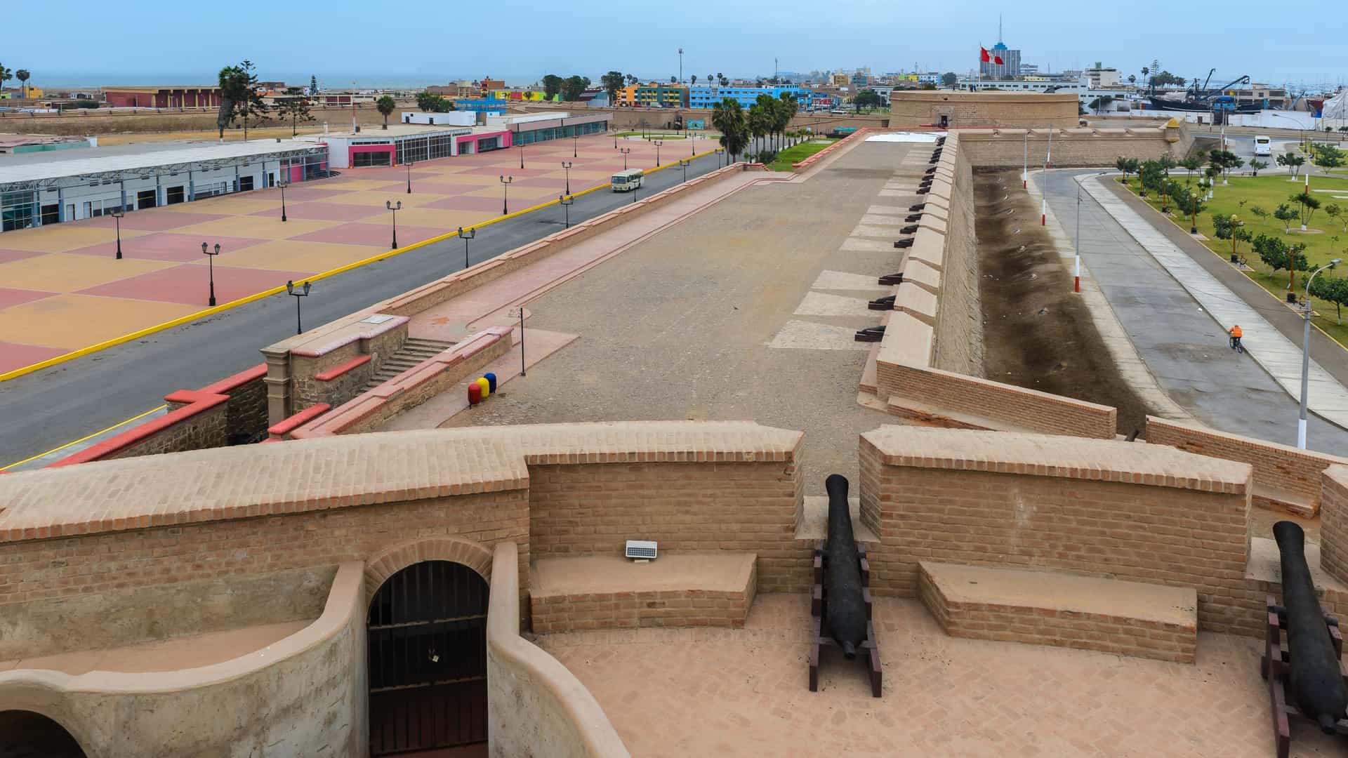 A ground-level view of the historic Real Felipe Fortress, a massive pentagonal stone structure in Callao, Peru. The image focuses on a circular bastion, the Torreón del Rey, with an old defensive cannon mounted on the rampart, aimed out over the green lawn toward the modern city buildings in the background under a cloudy sky, showcasing the fortress's role as a colonial-era coastal defense.