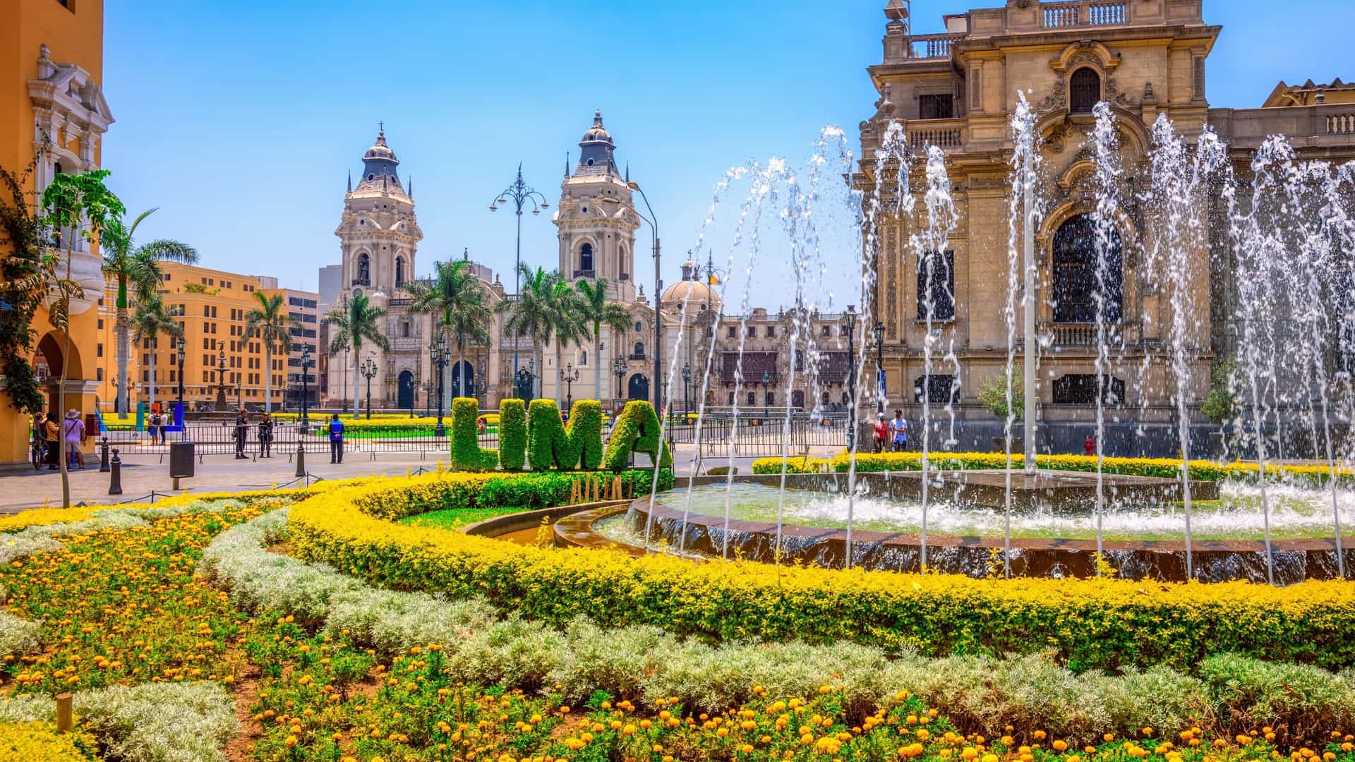 A wide-angle view of the historic Plaza de Armas (or Plaza Mayor) in Lima, Peru, featuring the ornate facade of the Lima Cathedral. The Baroque and Renaissance-style cathedral dominates the background, with the square's central bronze fountain and manicured green lawns in the foreground. The scene is framed by tall palm trees and other Spanish colonial-era buildings, highlighting the historical and architectural heart of the city.