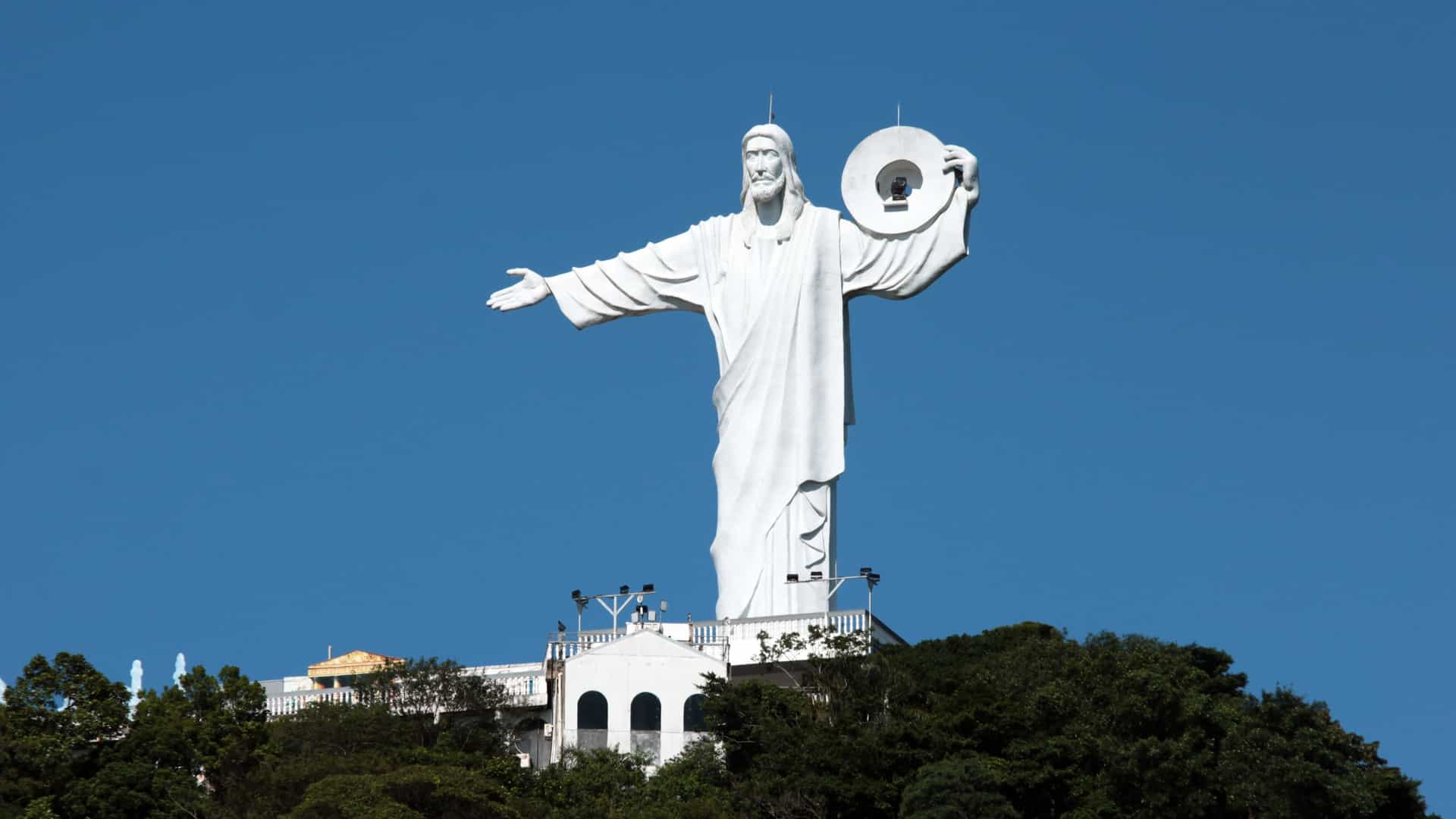 A nighttime long-exposure shot of the Cristo Luz (Christ Light) monument perched on a hill overlooking the city of Balneário Camboriú, Santa Catarina, Brazil. The statue is dramatically illuminated with multi-colored lights, creating streaks in the sky.