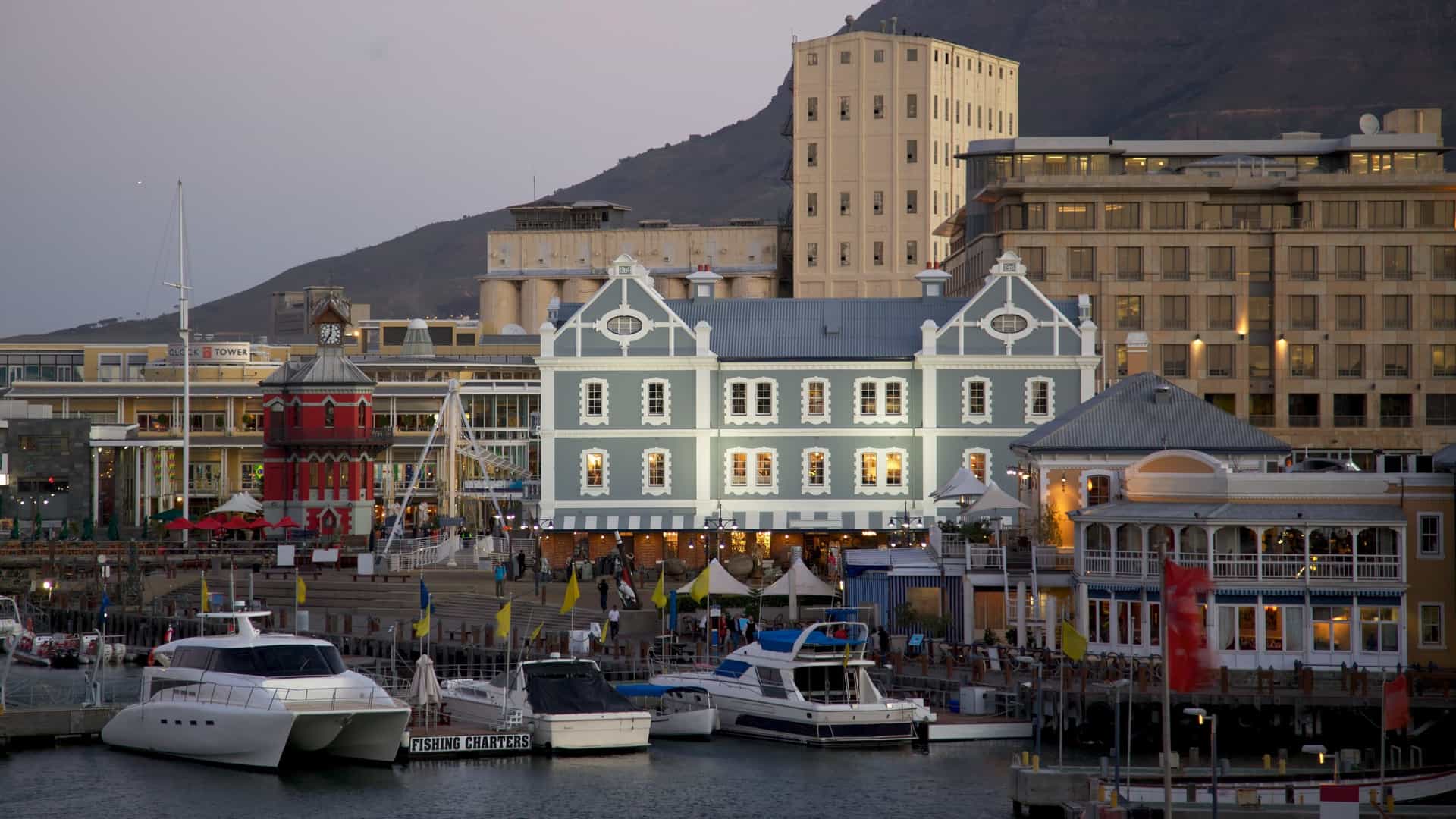  The Victoria & Alfred Waterfront in Cape Town, South Africa, with boats in the harbor and a variety of buildings along the waterfront. In the background, Table Mountain rises majestically, its flat top a prominent feature of the landscape.