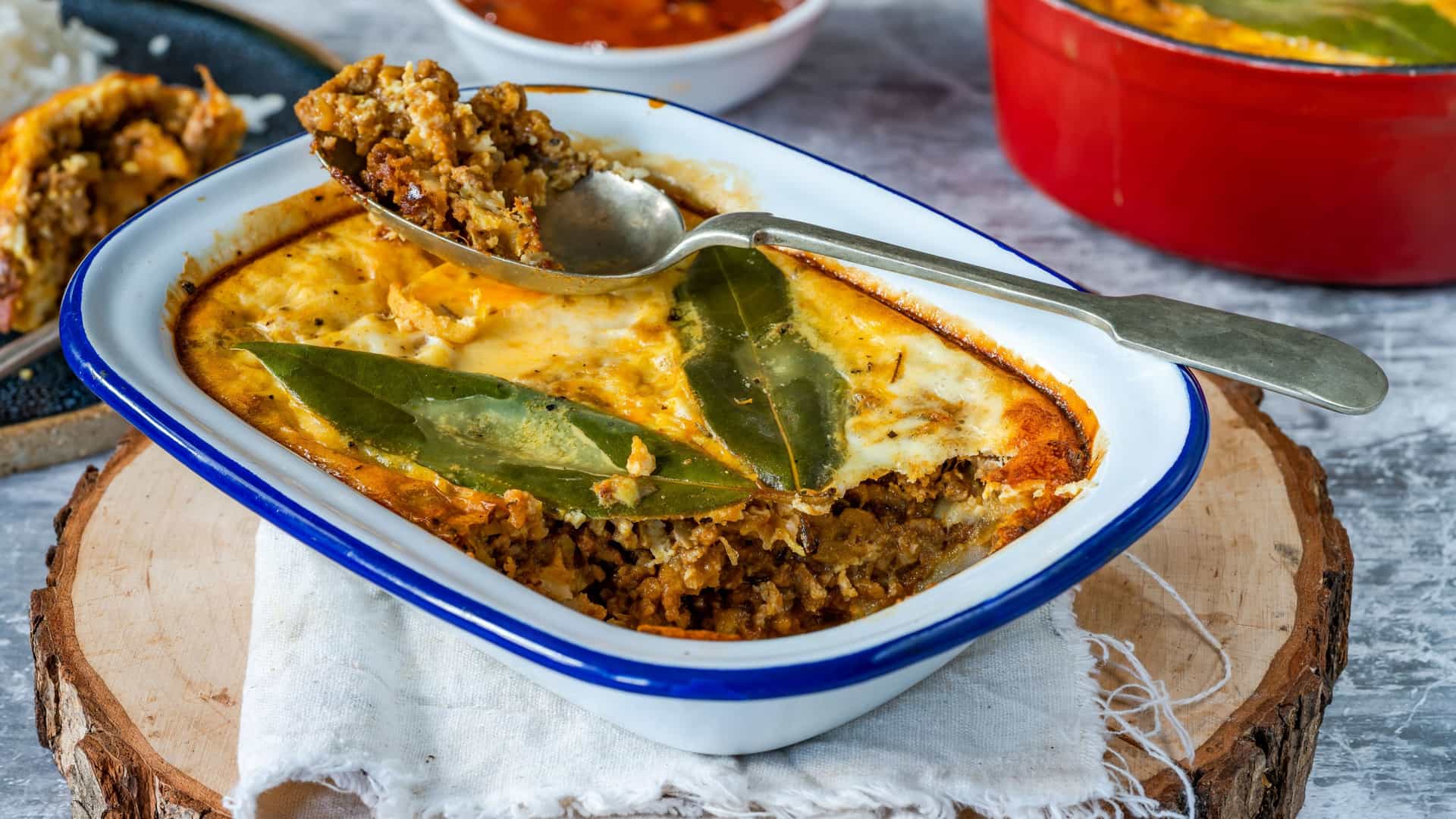 A close up shot of a baked bobotie, a South African dish consisting of a curried minced meat base and a golden brown egg custard topping. The surface of the dish is garnished with a few bay leaves.