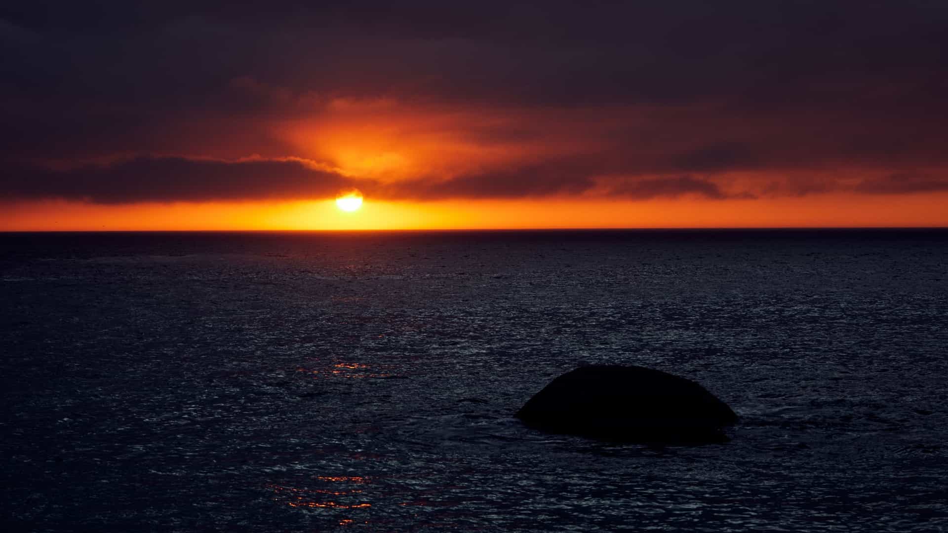  A serene cloudy sunset over Camps Bay, South Africa, with the sky ablaze in hues of orange and pink. The calm ocean reflects the vibrant colors, and the silhouettes of mountains are visible in the background.