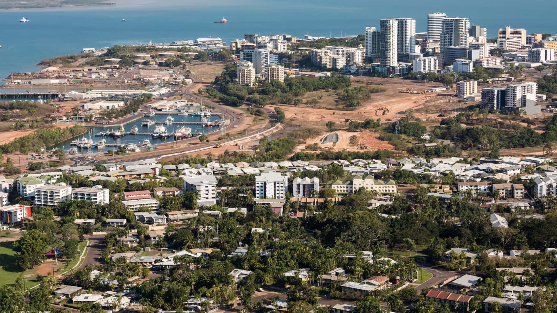 An aerial photograph of Darwin, the capital city of Australia's Northern Territory, showcasing the modern urban area, the curving coastline of the harbor, and the surrounding tropical environment.