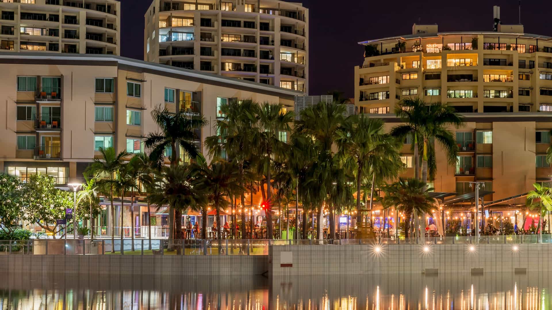  A night view capturing an outdoor dining experience, reminiscent of a Dinner at a Darwin Waterfront Restaurant. The photo shows a rustic wooden building built over dark water, illuminated by warm interior light, with patrons blurred as they dine. In the distance, the city skyline is reflected on the calm water under a cloudy night sky.