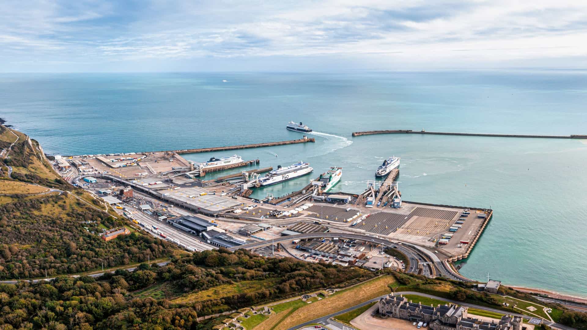  An aerial view of the Port of Dover in England. The image shows the busy harbor with several large ferries and other ships docked or navigating the water. The port infrastructure, including docks and roads, is visible, with the coastline and the town of Dover in the background.