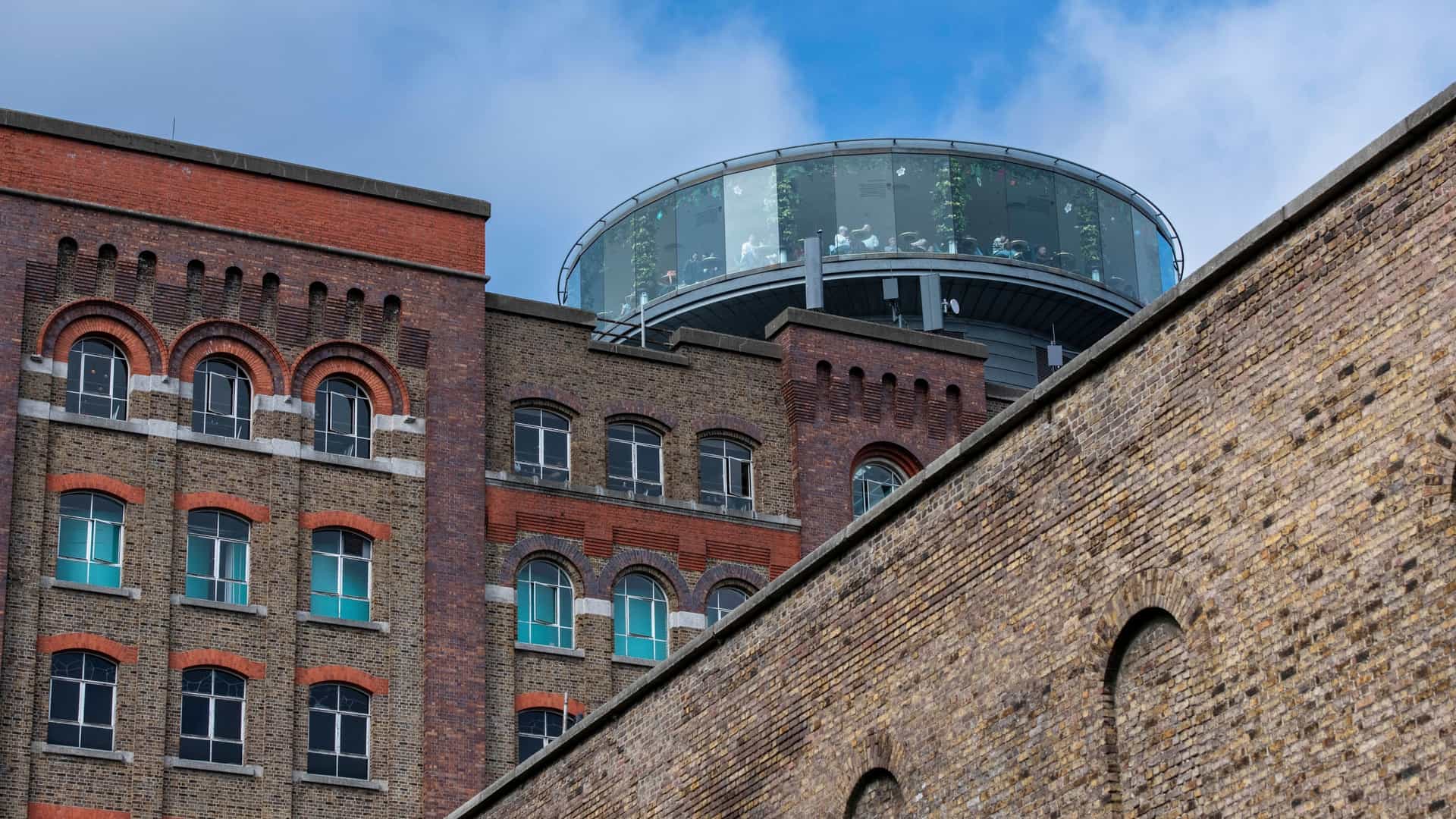 A large exterior view of the historic brick buildings of the Guinness Storehouse in Dublin, Ireland. The architecture features tall, multi-story facades with numerous windows and industrial-style stonework.