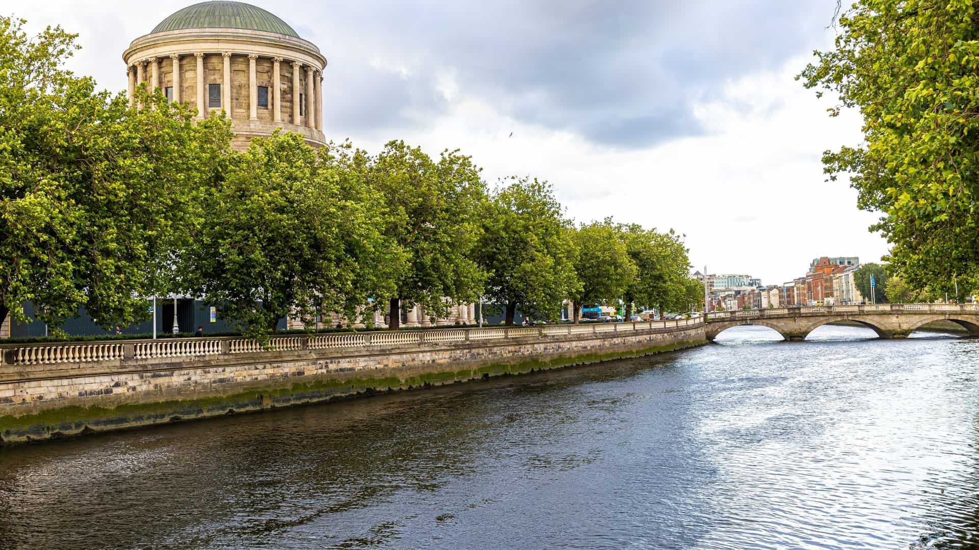  A view along the River Liffey in Dublin, Ireland, dominated by the neo classical dome of the Four Courts building on the left bank. A line of lush green trees runs along the stone quay, with an arched stone bridge visible in the background.