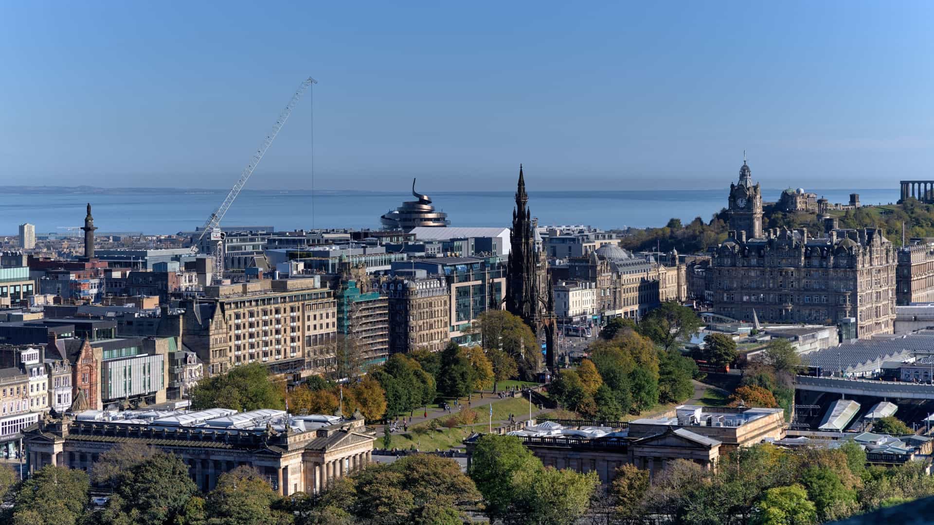 -A panoramic view of the historic Edinburgh cityscape, likely taken from an elevated vantage point like Calton Hill or Arthur's Seat. The shot features a dense collection of traditional Scottish architecture, church spires, and historic buildings spreading across the landscape, with the Firth of Forth estuary and hills visible in the distant background under a clear blue sky.