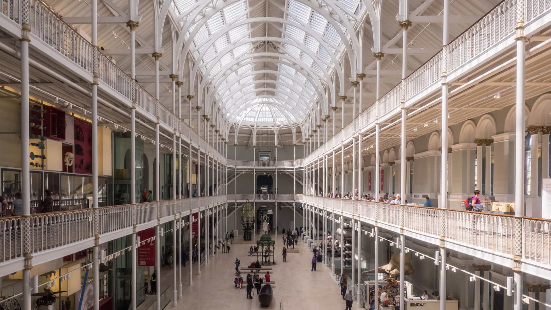 A high-angle interior view of the Grand Gallery or main atrium of the National Museum of Scotland in Edinburgh. The photograph captures the magnificent Victorian architecture, featuring multiple tiered balconies, ornate cast-iron railings, and an expansive glass ceiling that floods the exhibit space with natural light. The vast hall contains various displays and is busy with visitors exploring the cultural and historical collections.